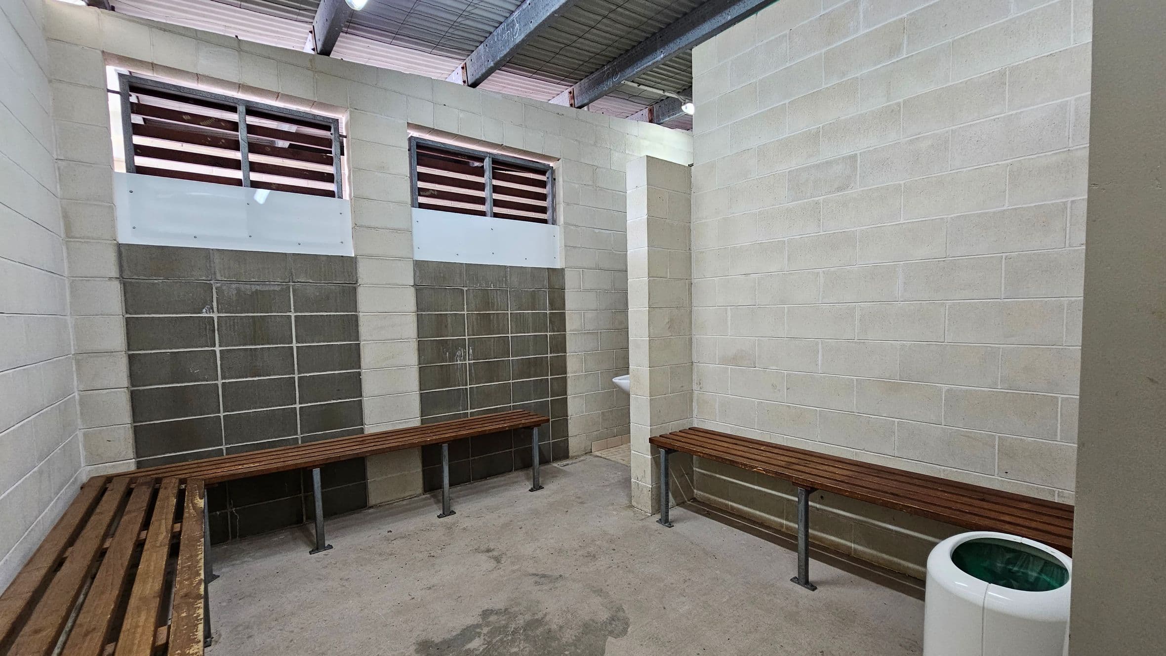 A room with concrete block walls and a corrugated metal ceiling. On the left and right, there are wooden benches attached to the walls. Two small windows with horizontal slats are on the left wall. In the corner, a small white sink is mounted on the wall. The floor is concrete with visible stains. A white cylindrical bin with a green interior is on the right side.