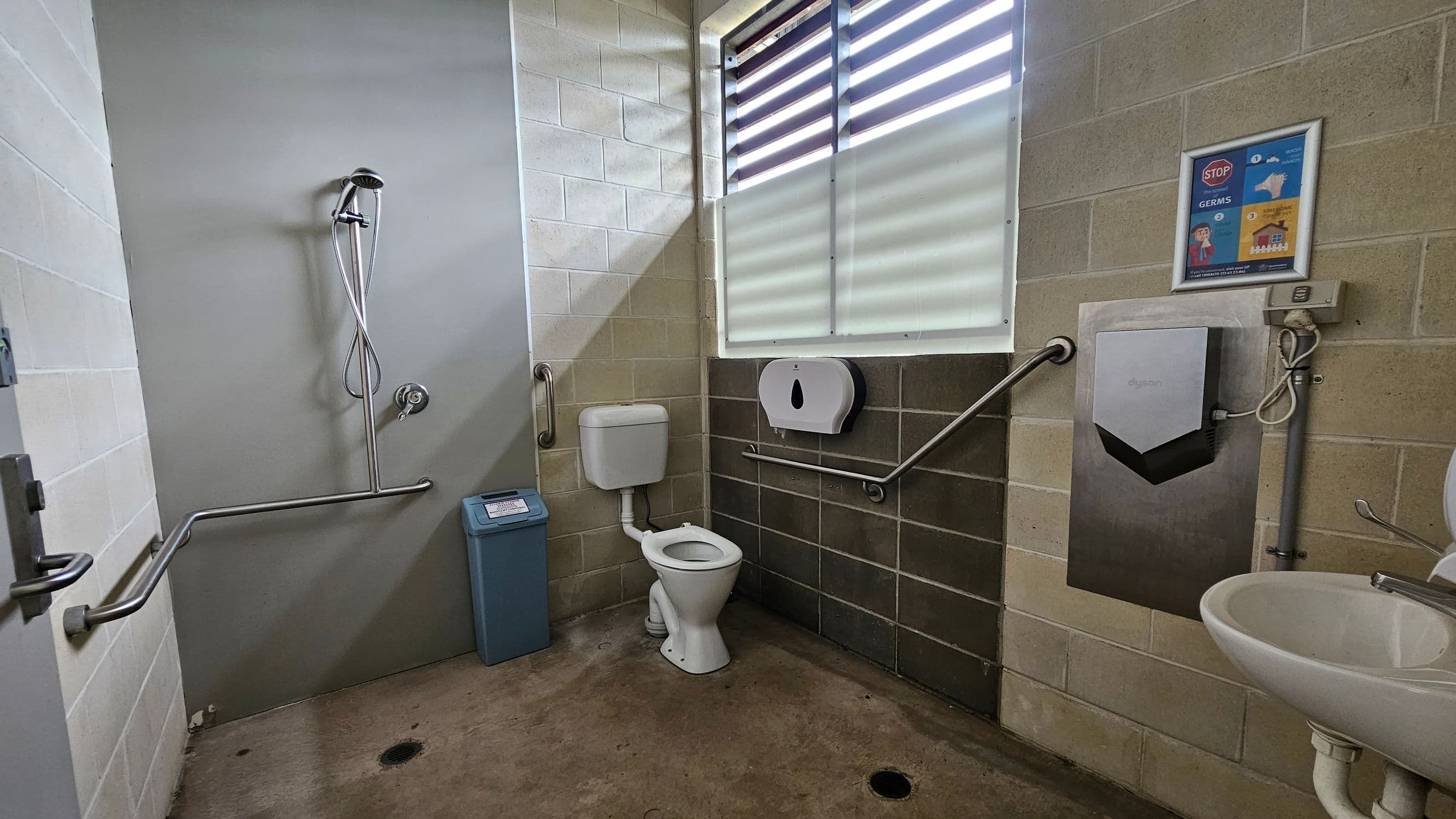 A public bathroom with a concrete floor and tiled walls. On the left, there's a shower with a handheld showerhead and a metal grab bar. In the centre, a toilet with a cistern is positioned next to a blue bin. On the right, a wall-mounted hand dryer and a paper towel dispenser are above a sink. A poster about hygiene is on the wall. The window has horizontal slats allowing light in.