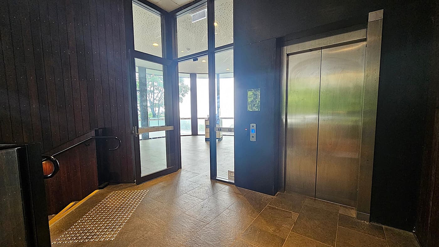 A modern interior with a lift on the right, featuring metallic doors and a control panel. To the left, a dark wooden wall with a handrail and a textured floor section. In the centre, a glass door leads to a bright area with large windows and visible trees outside. The floor is tiled, and the ceiling has recessed lighting.
