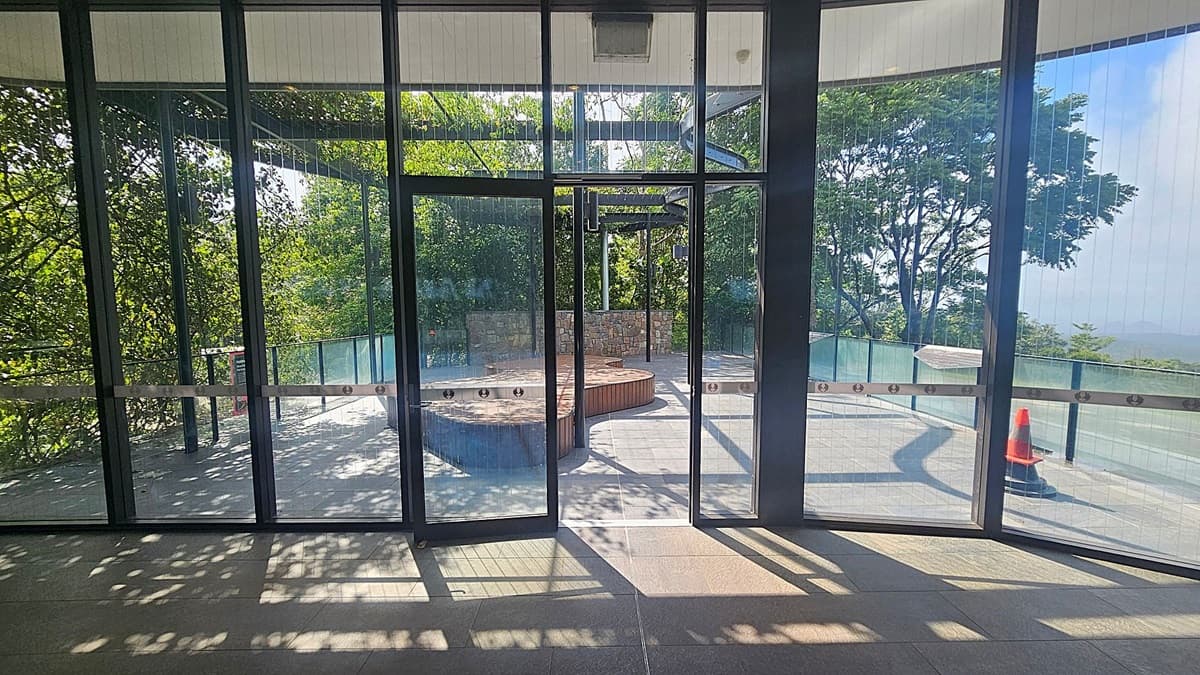 Glass doors open to a terrace with a circular wooden bench in the centre. The terrace is surrounded by a glass railing, and a single orange traffic cone is visible on the right. Lush green trees are in the background, casting shadows on the ground. The floor inside is tiled, and the ceiling is white.