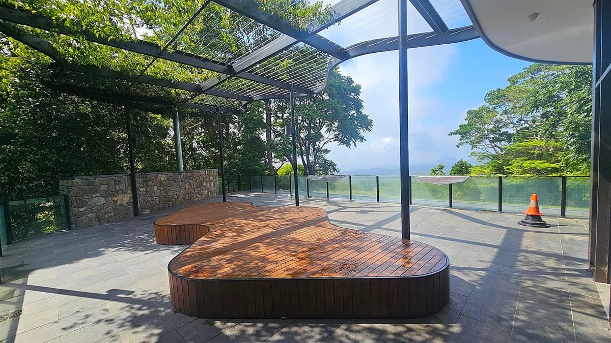 A terrace with a wooden bench in the centre, surrounded by a stone wall on the left and glass railings on the right. The floor is tiled, and a metal pergola overhead is partially covered with greenery. In the background, there are trees and a view of the sky. A single orange traffic cone is placed near the glass railing on the right.