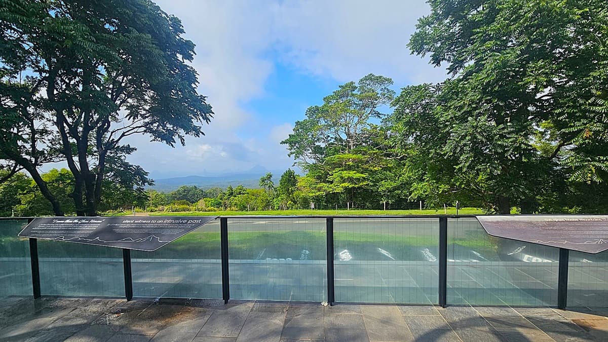 A scenic view with a glass railing in the foreground, featuring informational panels. Beyond the railing, a lush landscape with dense green trees on both sides frames the view. In the middle distance, a road runs horizontally, and further back, more greenery extends towards a hazy horizon. The ground is paved with dark stone tiles.