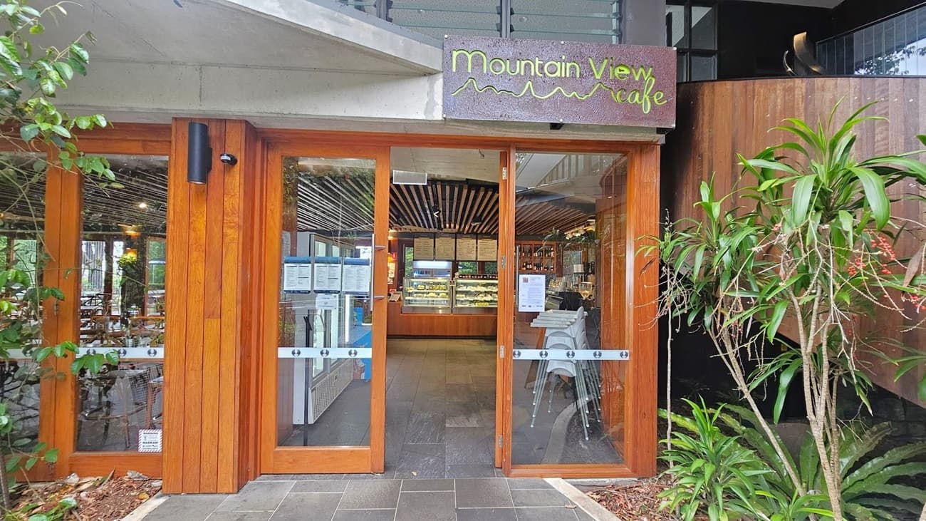 Entrance to a café with wooden-framed glass doors. Above the doors, a sign reads "Mountain View Café" in green lettering. Inside, there are tables and chairs, and a display counter with food items. The ceiling has a striped wooden design. On the left, there are green plants climbing the wall, and on the right, potted plants with long leaves and red berries. The floor is tiled in dark grey.
