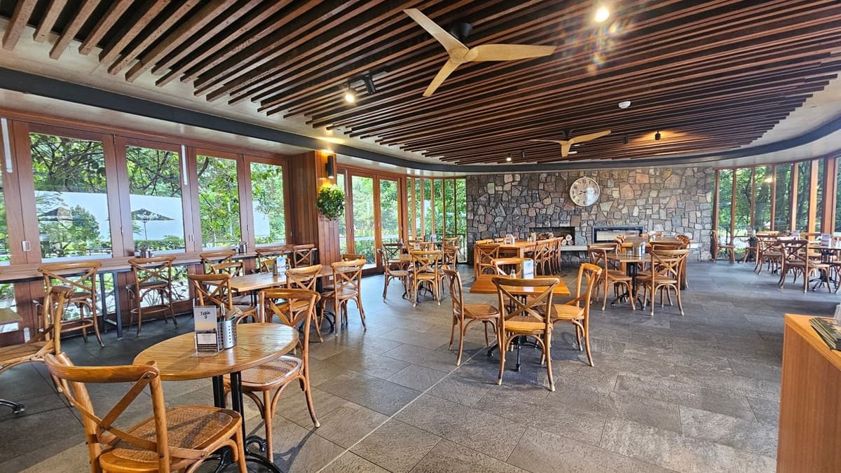 A spacious restaurant interior with wooden tables and chairs arranged neatly. Large windows on the left offer views of greenery outside. The back wall is made of stone with a large clock and a fireplace. The ceiling features wooden slats and ceiling fans. The floor is tiled in a grey stone pattern.