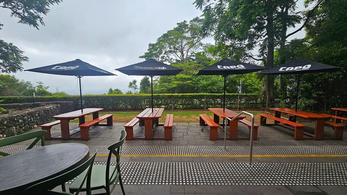 Wooden picnic tables with black umbrellas are arranged on a stone patio. In the foreground, a round table with chairs is visible. The background features a low stone wall, a hedge, and tall trees. The ground is paved with textured tiles, and a metal handrail is present.