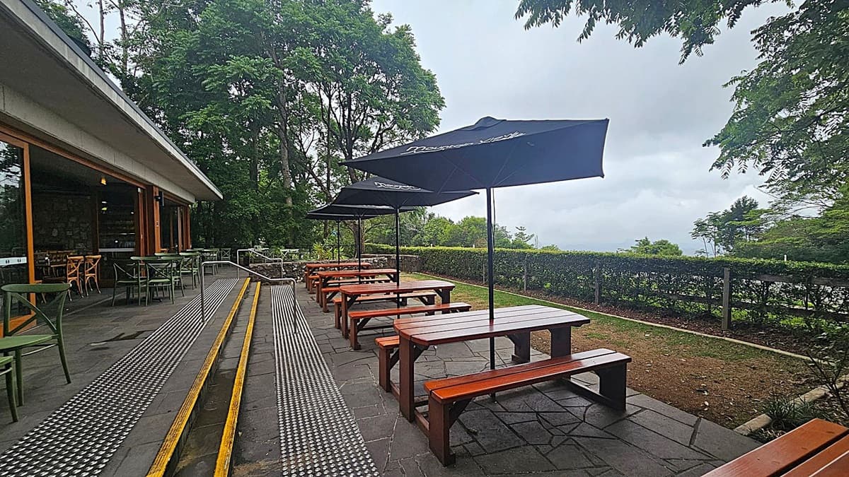 Outdoor seating area with wooden picnic tables and large black umbrellas on the right. A paved walkway with yellow-striped ramps runs through the middle. On the left, there is a building with large windows and green chairs and tables. The background features lush green trees and a hedge, with a view of the landscape beyond.