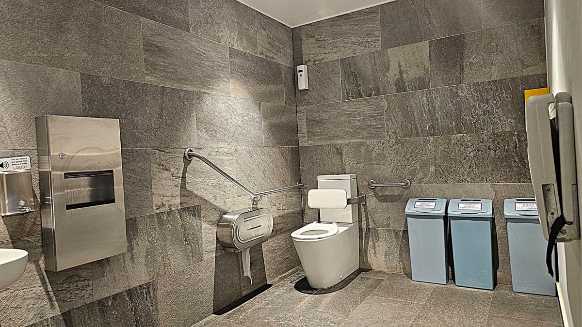 A public toilet with grey stone walls and floor. On the left, there's a stainless steel hand dryer and a paper towel dispenser. In the centre, a toilet with a white seat and a support bar on each side. Above the toilet, a small white air freshener is mounted on the wall. On the right, three blue bins with labels for different types of waste. The ceiling is white with recessed lighting.