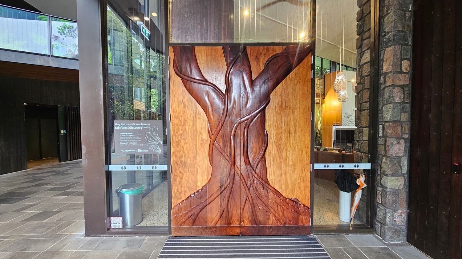 A large wooden door with an intricate tree carving stands in the centre. To the left, a glass panel displays information about the Mary Cairncross Scenic Reserve Rainforest Discovery Centre. Below the panel is a silver bin. To the right, another glass panel shows the interior with hanging lights and a stone wall. The floor is tiled with grey and black stripes.