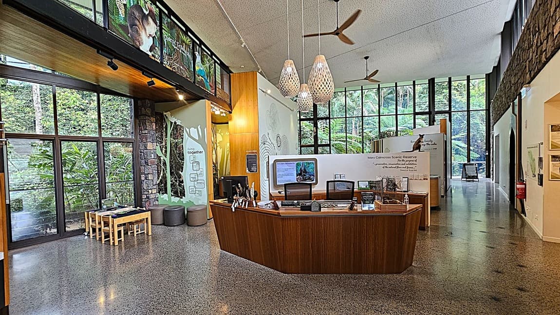 A spacious interior with a polished concrete floor and high ceiling. On the left, large windows reveal lush greenery outside. Below the windows, a small wooden table with chairs is set up. In the centre, a wooden reception desk displays informational materials. Above, three pendant lights hang from the ceiling. The right side features more large windows and a stone wall, with informational displays about the Mary Cairncross Scenic Reserve.