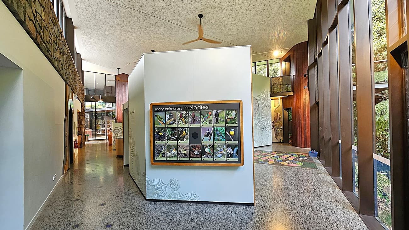 Interior of a modern building with a polished concrete floor. In the centre, a display board titled "Mary Cairncross Melodies" features images of birds. The left side has a stone wall and large windows, while the right side has tall wooden panels and more windows overlooking greenery. The ceiling is white with a fan. In the background, there are more displays and a glass entrance.