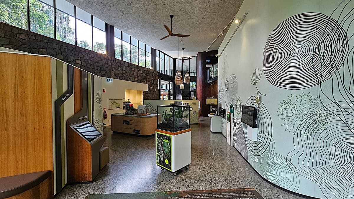 Interior of a modern exhibition space with a polished stone floor. On the left, there are wooden panels with interactive screens. In the centre, a glass display case on wheels contains a small exhibit. The right wall features artistic circular patterns and mounted screens. Large windows in the background allow natural light to illuminate the room. The ceiling has a fan and pendant lights, and a stone wall runs along the upper section.