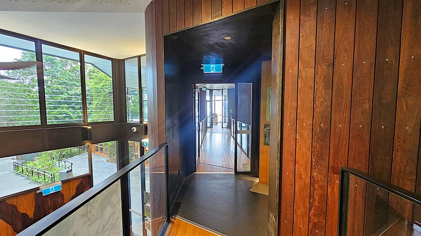 A corridor with wooden walls and a wooden floor leads to a glass door at the end. On the left, large windows with horizontal slats offer a view of greenery outside. The ceiling is light-coloured, and there are emergency exit signs above the doors. A glass railing runs along the left side of the corridor.