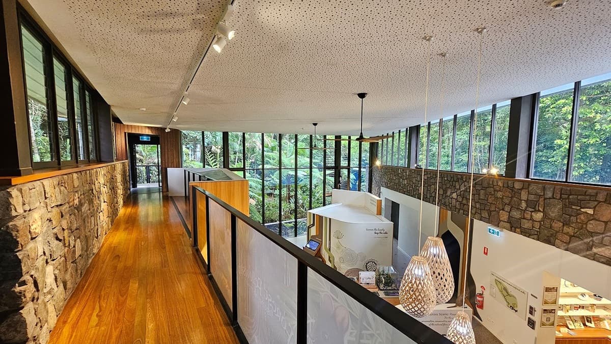 A modern interior with a wooden floor and a stone wall on the left. Large windows line the right side, offering a view of lush greenery outside. The ceiling is white with small perforations and features hanging lights. Below, a display area with informational panels and a map is visible. The space is well-lit with natural light streaming in through the windows.