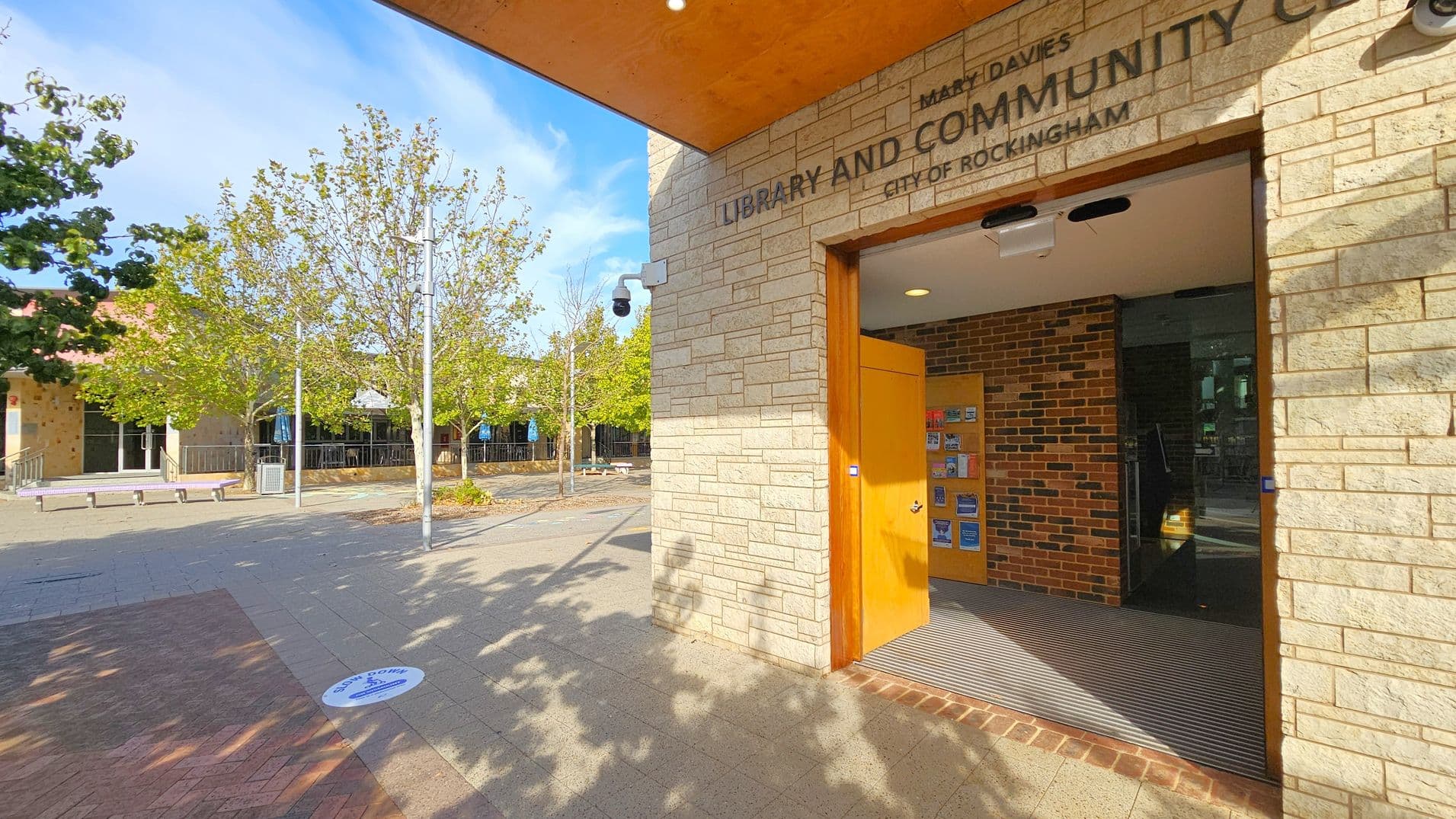 Entrance to a library and community centre with a stone facade on the right. The door is open, revealing a brick interior wall and a display of brochures. A security camera is mounted above. On the left, a paved area with trees and benches is visible. The ground has a circular social distancing marker. The ceiling above the entrance is orange.