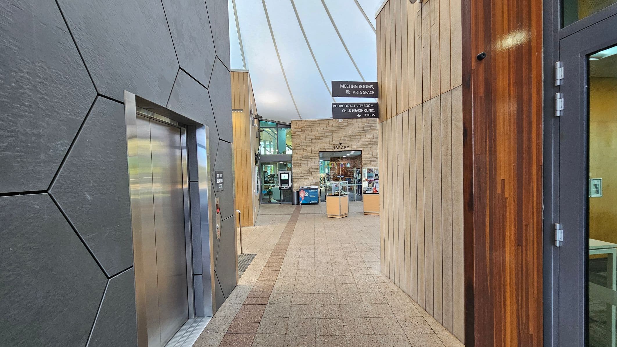 A modern indoor hallway with a geometric-patterned wall on the left featuring a lift. The floor is tiled with a central strip leading towards a library entrance in the background. On the right, there is a wooden-panelled wall and a glass door. Overhead, a curved ceiling with white panels and metal beams. Signs above indicate directions to meeting rooms, an arts space, a child health clinic, and toilets.