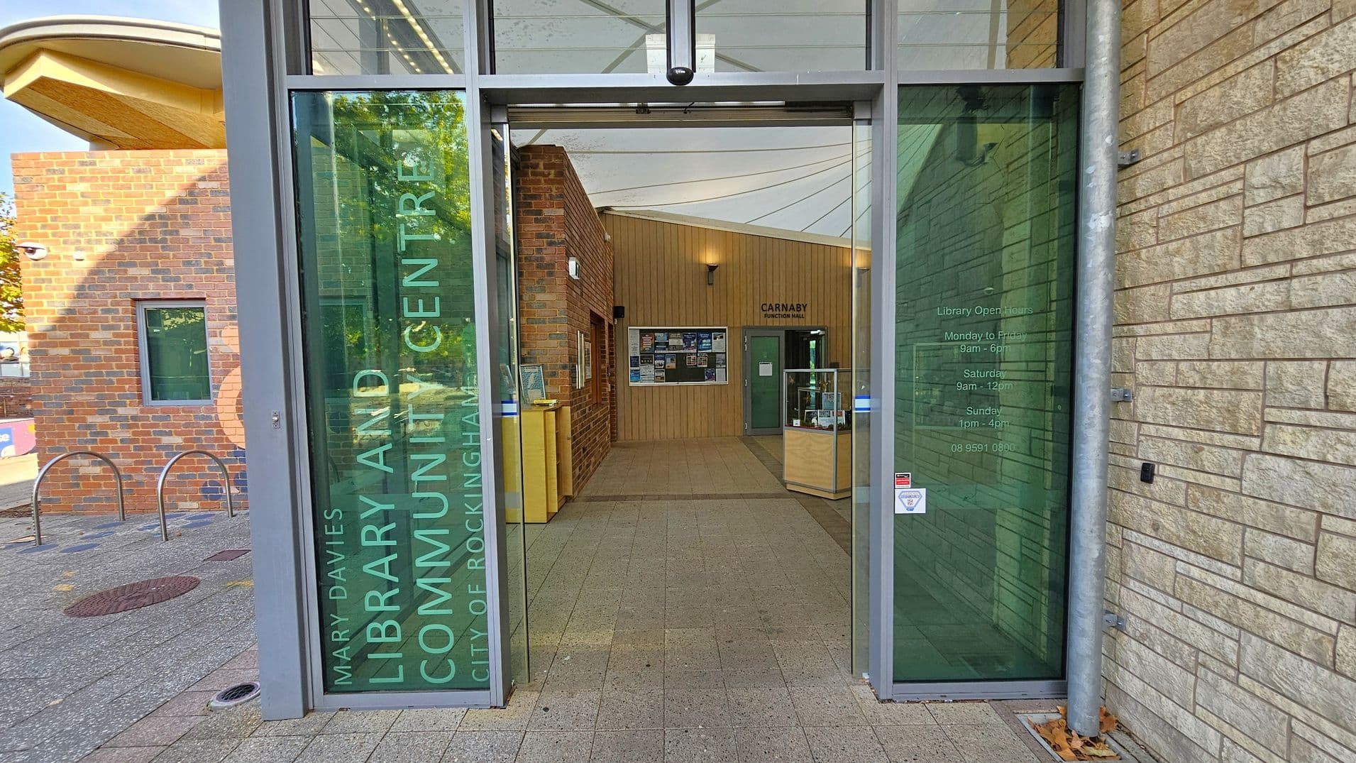 Entrance to a building with glass doors. The left door has text reading "Mary Davies Library and Community Centre, City of Rockingham." The right door displays library opening hours. Inside, a hallway with brick and wood-panelled walls leads to a notice board and a door marked "Carnaby Function Room." Display cases are on either side. The exterior features brick and stone walls, with a bicycle rack visible on the left. The floor is tiled, and the ceiling inside is white with lighting.