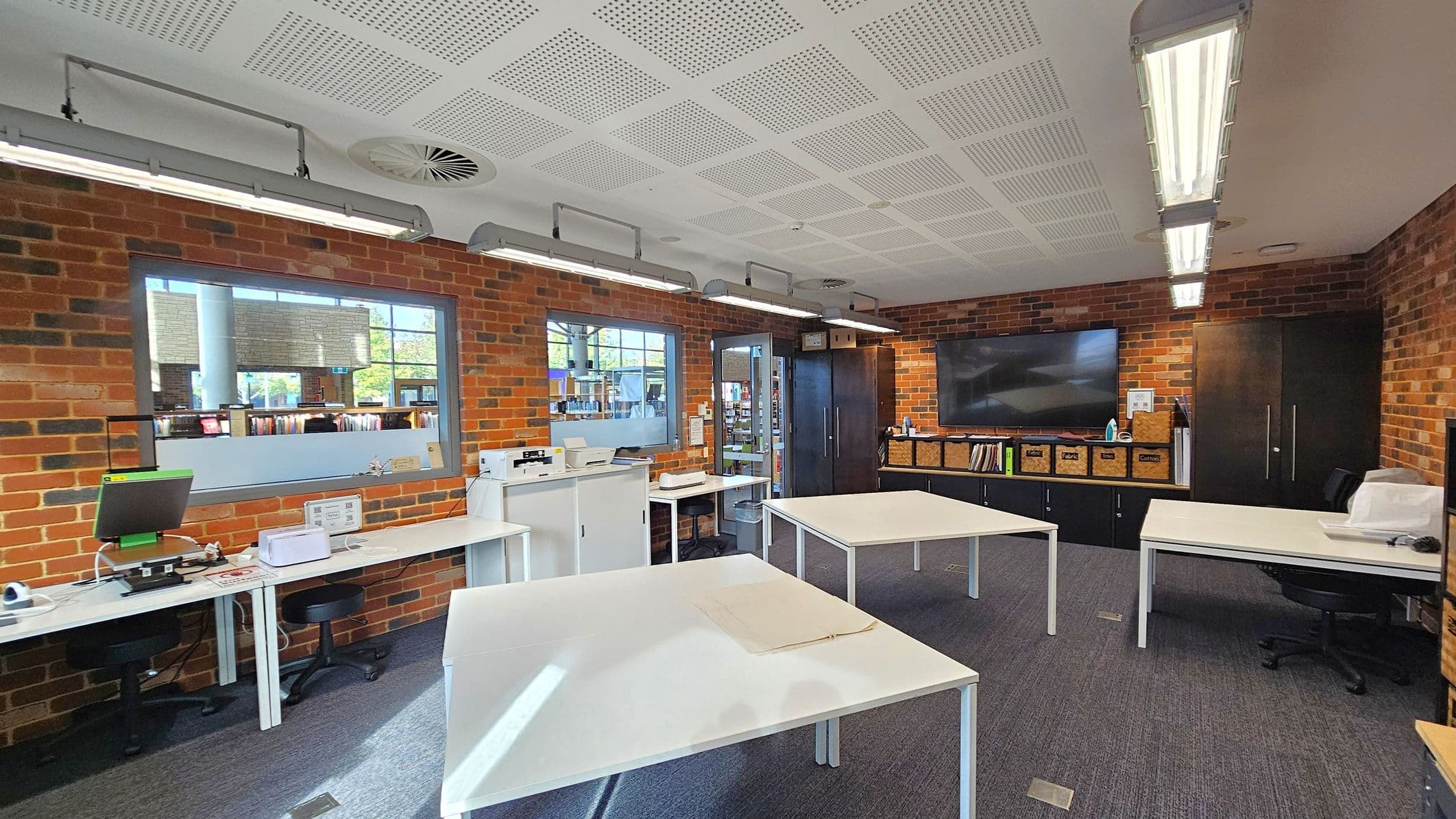 A room with brick walls and a perforated white ceiling. On the left, there are desks with computers and office equipment. In the centre, several white tables are arranged with a clear workspace. The right side features a large flat-screen TV mounted on the wall above a row of cabinets with labelled storage boxes. Fluorescent lights are suspended from the ceiling, and windows provide a view into another room with bookshelves. The floor is carpeted in grey.