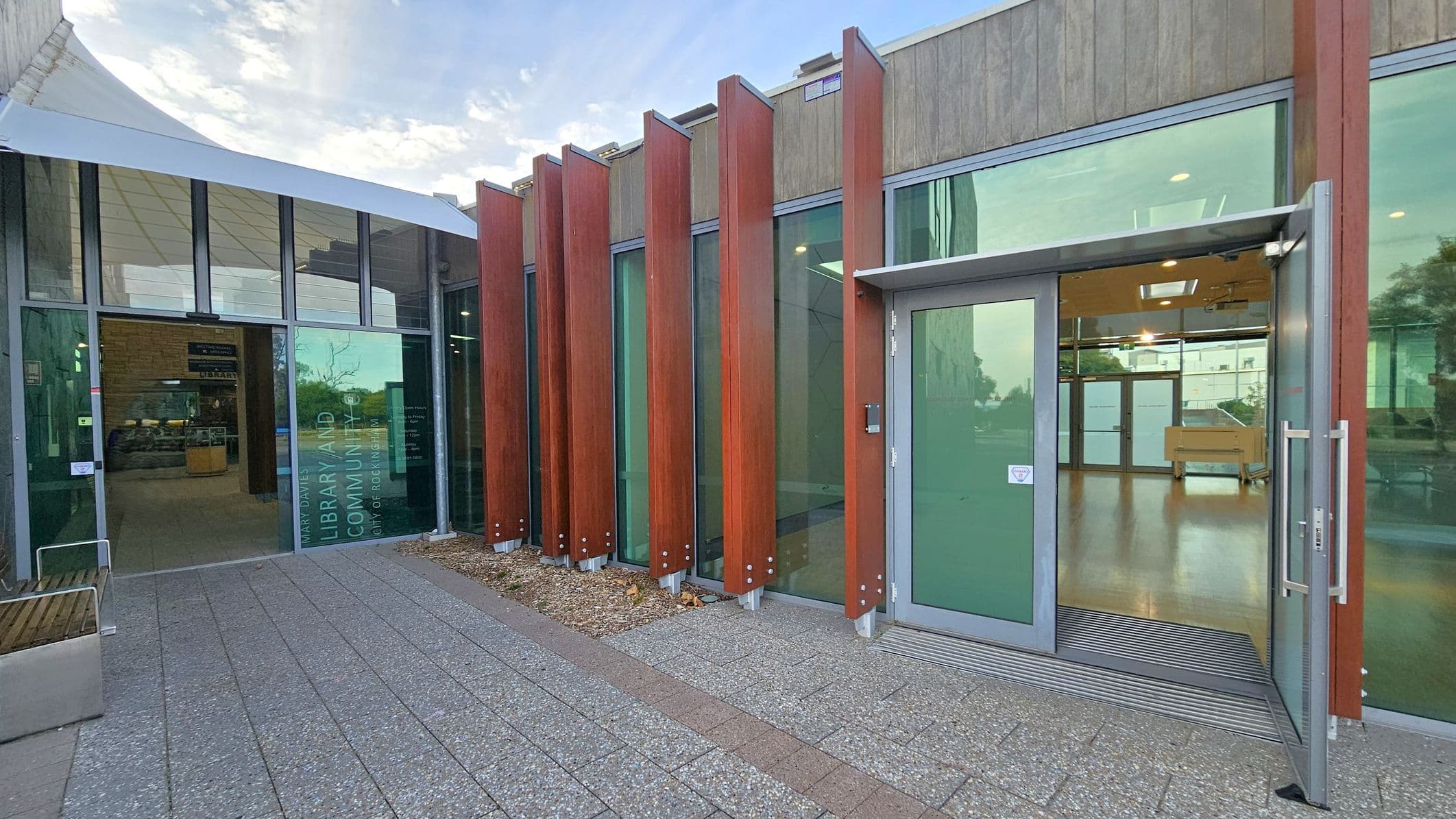 A modern building entrance with large glass panels and vertical wooden slats on the right. The door is open, revealing a spacious interior with wooden flooring. On the left, a glass facade with text indicating a library and community centre. The floor outside is paved with textured tiles, and a white canopy extends over part of the entrance.