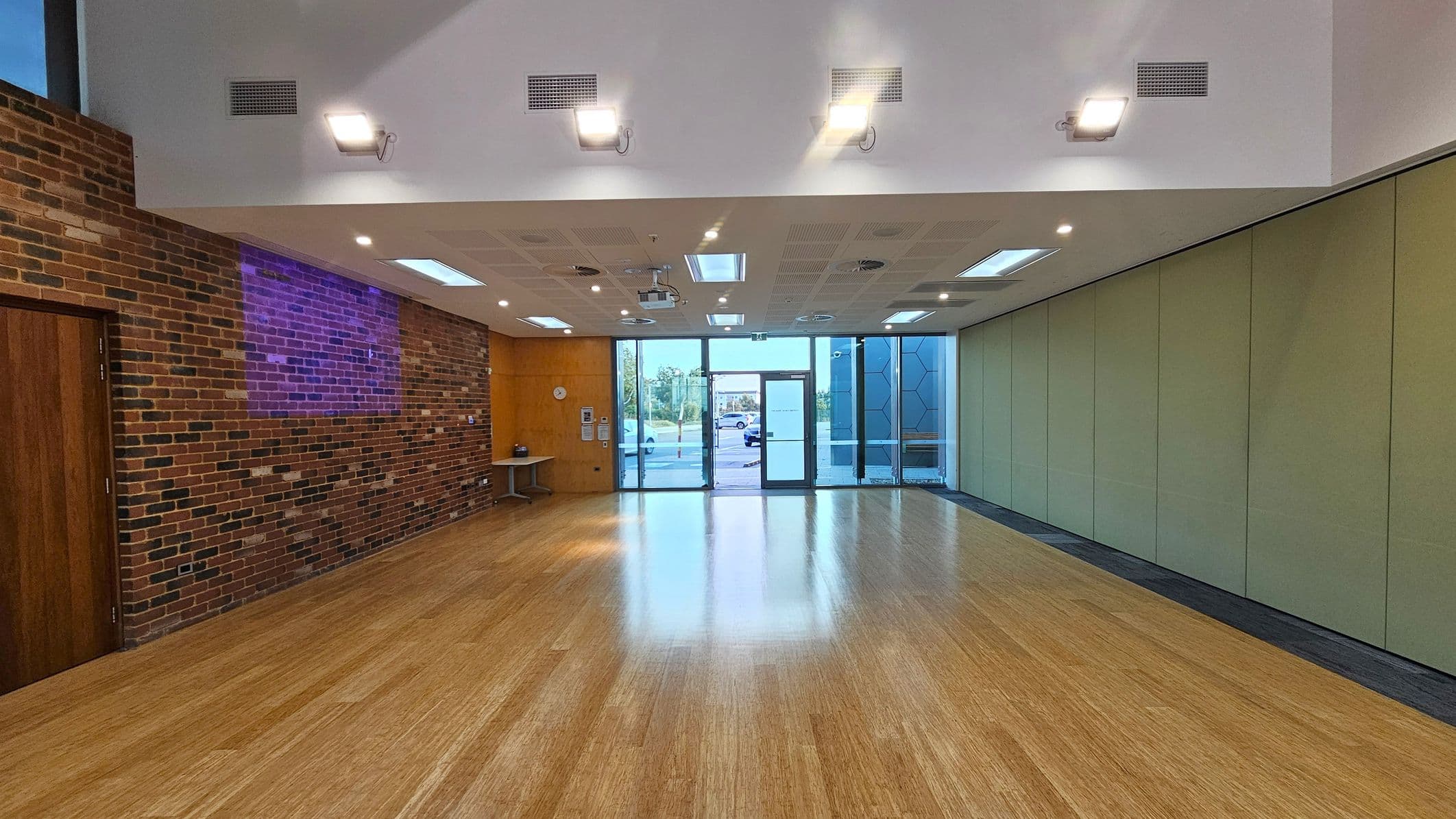 A spacious room with a wooden floor and a high ceiling featuring recessed lighting. On the left, there's a brick wall with a wooden door and a small table. The right side has a series of greenish partition panels. The back wall is mostly glass, with large windows and a central glass door leading outside. The ceiling has multiple light fixtures and ventilation grilles.