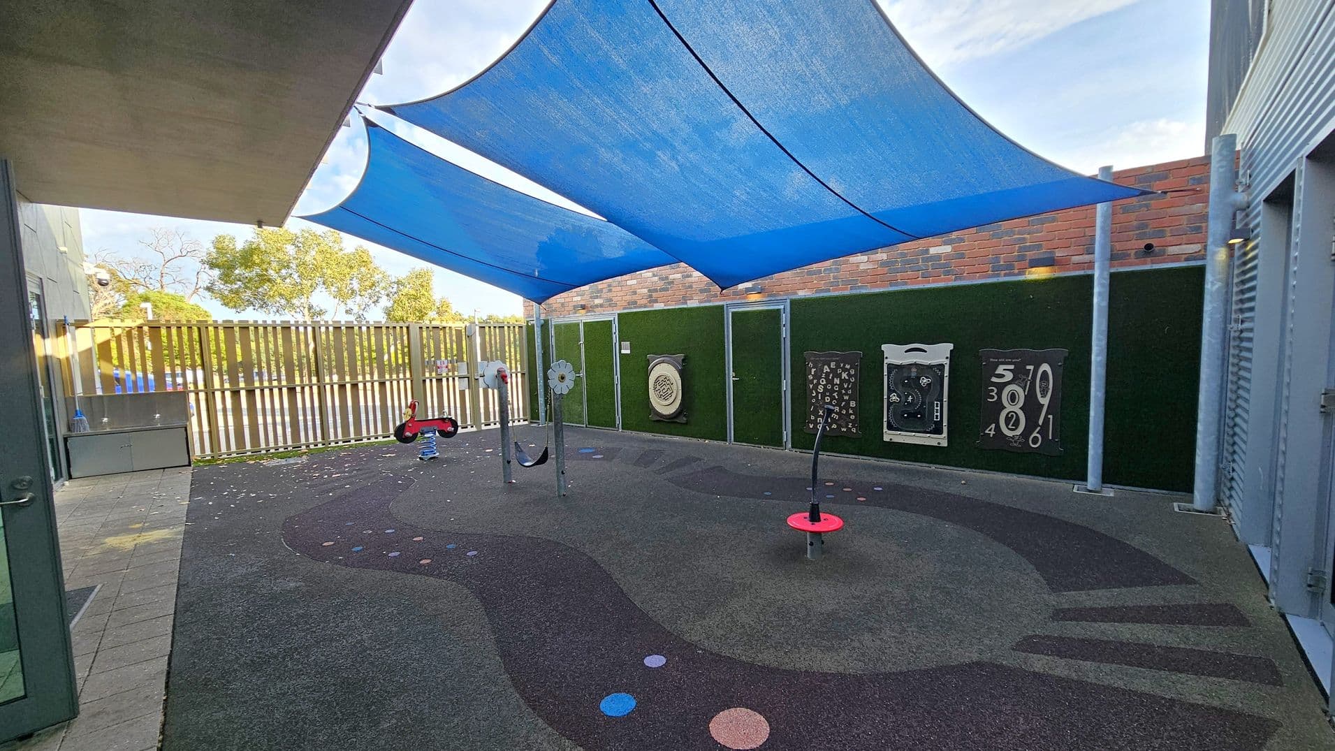 A children's play area with a soft, dark rubber floor featuring colourful circular patterns. In the centre, there are play structures, including a red spring rider and a spinning element. The area is shaded by large blue fabric canopies. The background has a green wall with interactive panels and a brick wall above. On the left, there's a wooden fence with trees visible beyond it. The ceiling is formed by the blue canopies, and the floor is a mix of dark and light grey rubber.