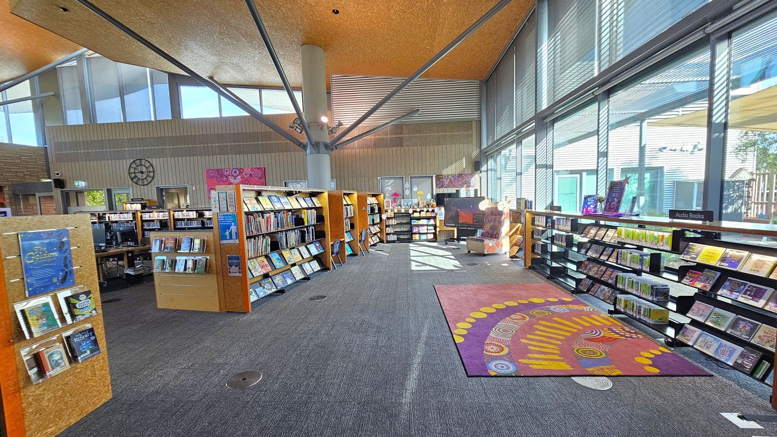 A library interior with a modern design. On the left, there are bookshelves filled with books and a display stand with posters. In the centre, more bookshelves are arranged in a semi-circle, with a large pillar supporting the ceiling. On the right, a long shelf holds audio books, and large windows let in natural light. The floor is carpeted with a colourful patterned rug in the middle. The ceiling is high with exposed beams, and there are artworks and a clock on the walls.