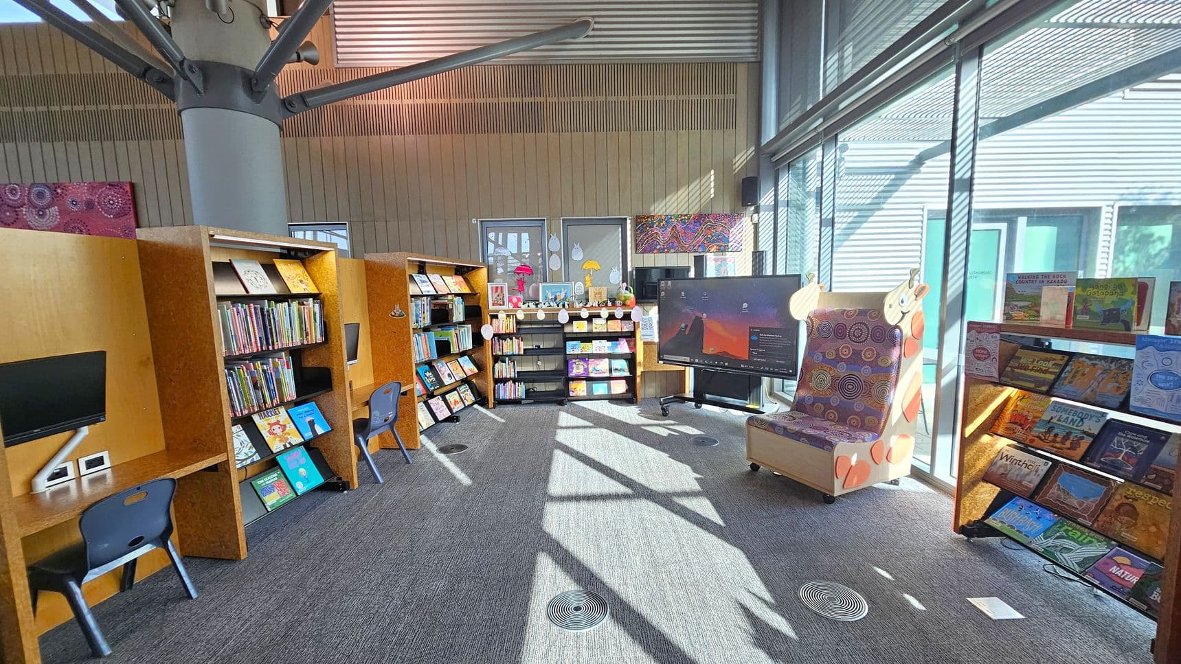 A library space with bookshelves filled with children's books on the left and centre. Two computer stations with blue chairs are on the left. In the centre, a small black bookshelf holds more books and decorations. A large digital screen is on the right, next to a colourful giraffe-themed chair. The right side features a window with blinds, allowing sunlight to illuminate the room. The floor is carpeted, and the ceiling has exposed beams and a large column.