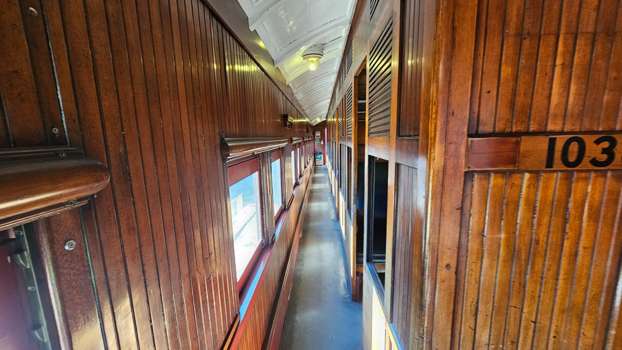 A narrow corridor inside a vintage train carriage with wooden panelling on both sides. On the left, large windows allow natural light to illuminate the space. The right side features numbered compartments, with "103" visible. The floor is a smooth, dark material, and the ceiling is white with a central light fixture. The overall atmosphere is warm and nostalgic, with a focus on the rich wood tones.