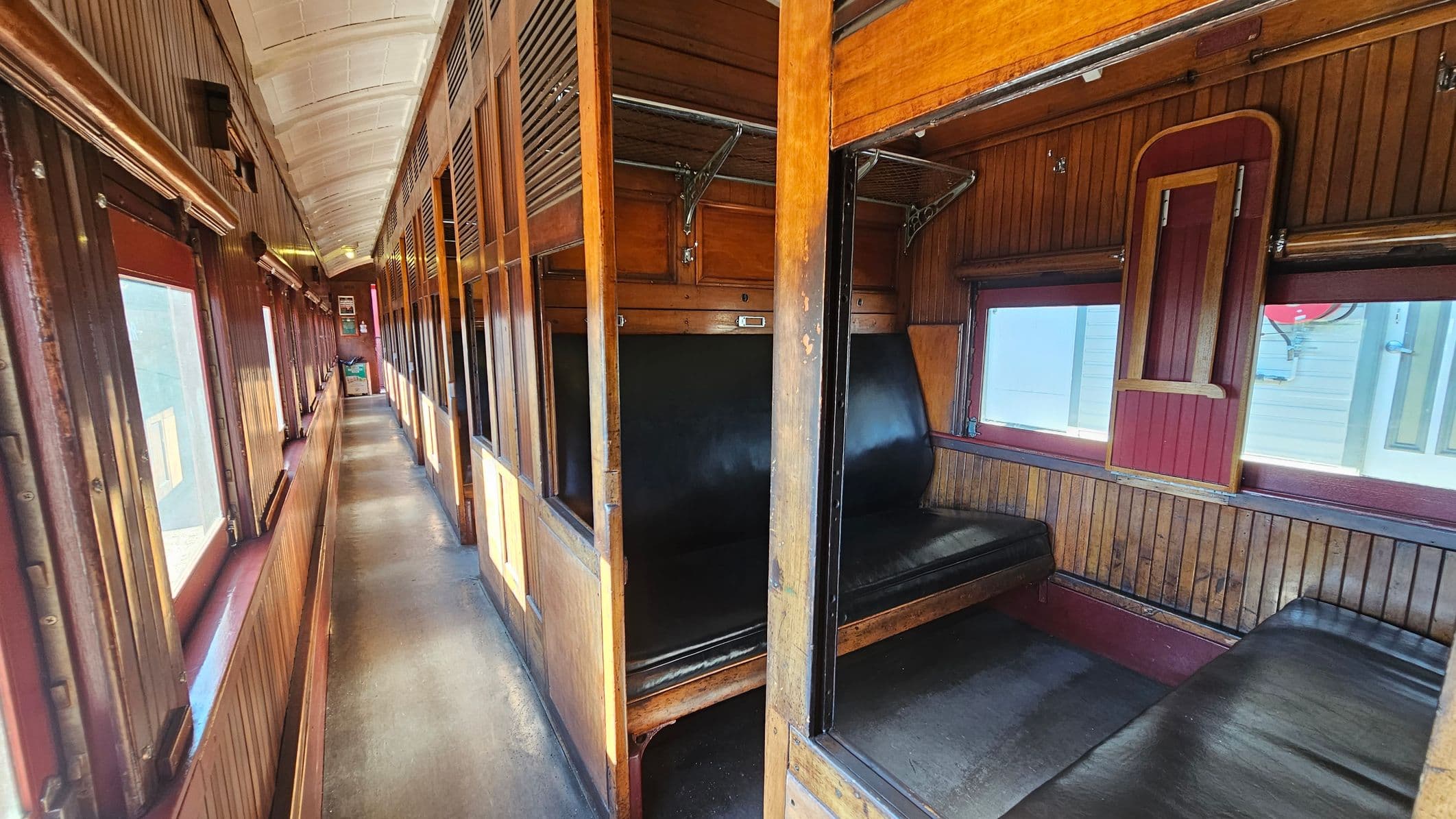 A vintage train carriage interior with wooden panelling. On the right, there are black leather bench seats in a compartment with a small window and a foldable wooden table. The left side features a narrow corridor with large windows, allowing light to stream in. Overhead, there are luggage racks. The ceiling is arched and painted white, contrasting with the dark wood. The floor is a simple, dark material.