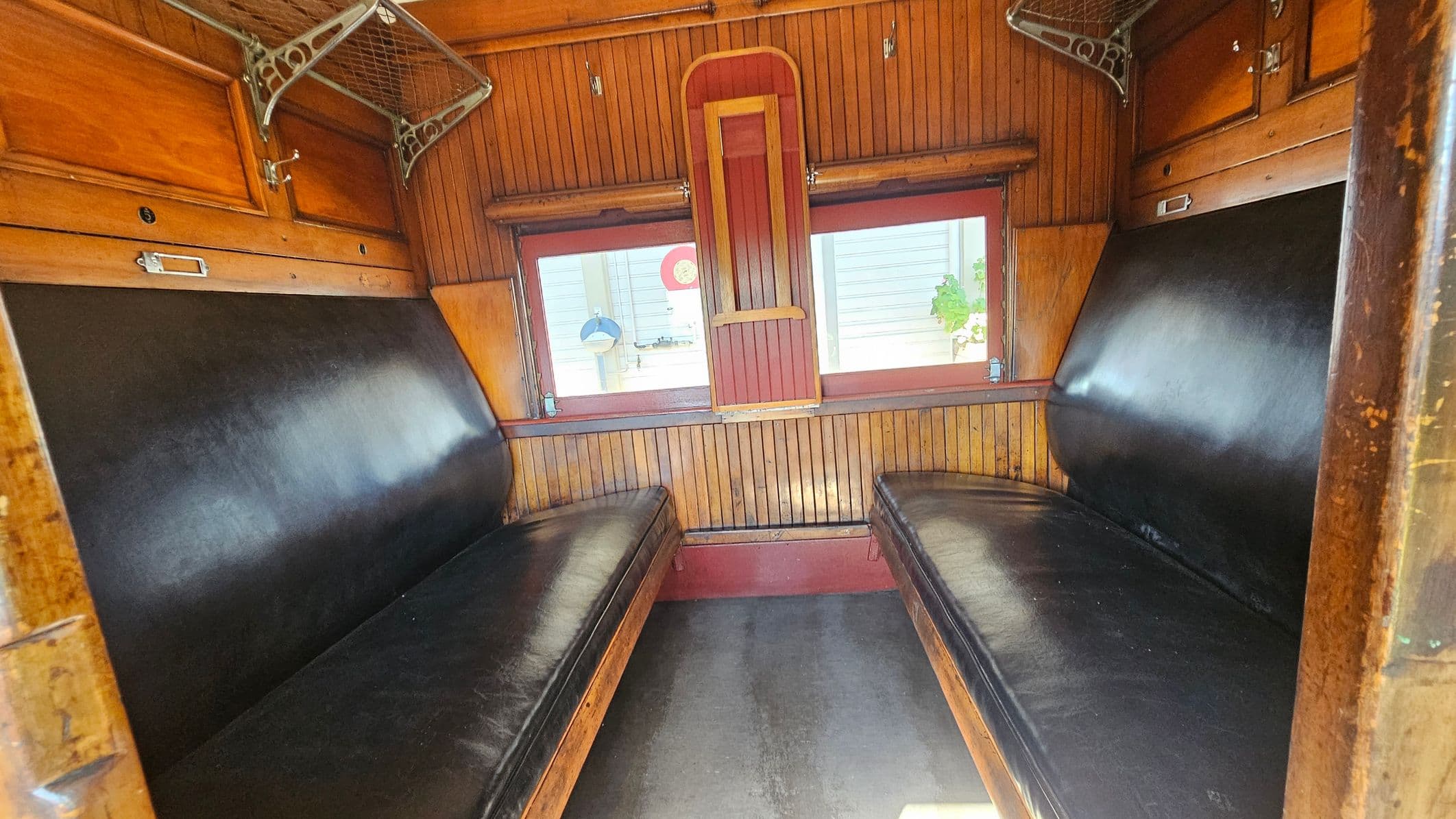 Interior of a vintage train compartment with two black leather benches facing each other. The walls and ceiling are lined with wood panelling. Above each bench, there is a metal luggage rack with a net design. The centre features a window with a wooden frame, partially covered by a red and wood panel. The floor is dark and smooth, contrasting with the warm wood tones.
