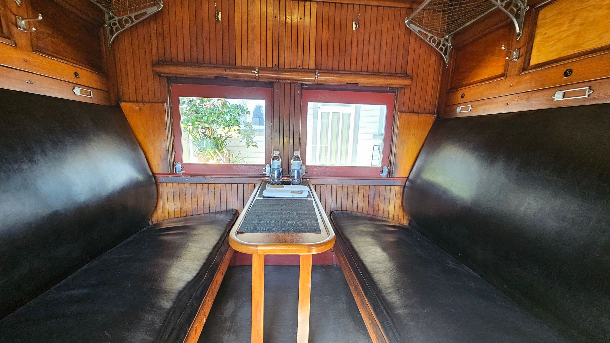 A vintage train compartment with wooden panelling and two black leather benches facing each other. In the centre, a narrow wooden table holds two bottles of water and a small tray with cutlery. Above the benches, metal luggage racks are visible. The windows in the background reveal a view of greenery and a building exterior. The ceiling is wooden, matching the walls, and the floor appears to be dark.
