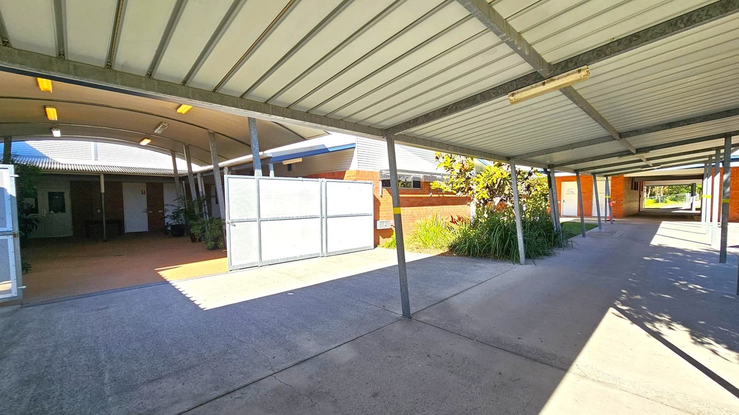 A covered walkway with a metal roof supported by metal beams. On the left, there is an entrance to a building with a curved roof and white panels. The building has brick walls and some plants near the entrance. On the right, a pathway leads to another section of the building, with more brick walls and greenery. The floor is concrete, and there are shadows cast by the structure and surrounding plants.