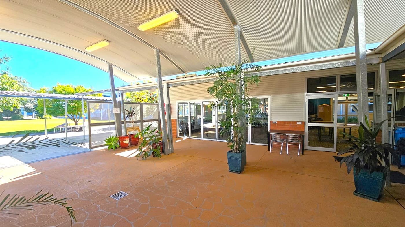 A covered outdoor area with a curved metal roof and fluorescent lights. The floor is a patterned terracotta-coloured surface. In the centre, there are potted plants, including a tall one in a square pot. On the right, glass doors lead inside, with metal chairs nearby. The left side opens to a grassy area with trees, visible through glass panels and a metal frame.