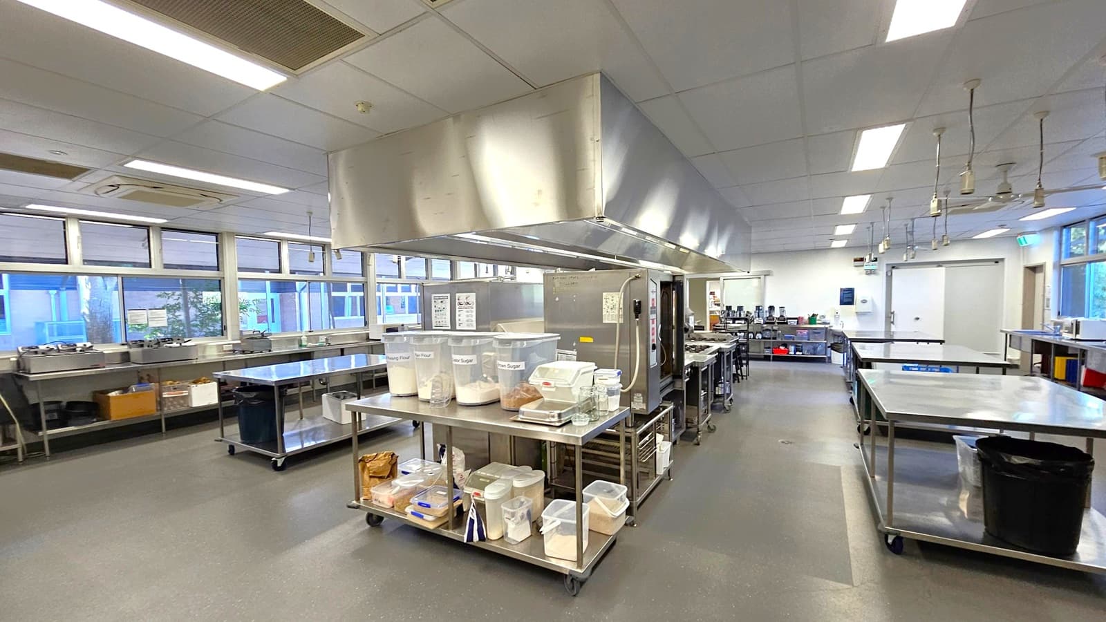 A spacious commercial kitchen with stainless steel worktables in the centre, holding large containers of ingredients like flour and sugar. On the left, there are windows and additional workstations with various kitchen equipment. The right side features more tables and a large refrigerator. The ceiling has fluorescent lighting and ventilation systems, while the floor is a smooth, dark material. The background includes more kitchen equipment and storage areas.