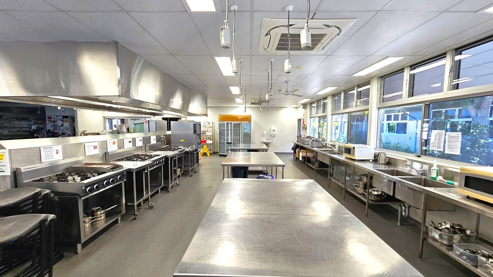 A commercial kitchen with stainless steel worktops in the centre. On the left, there are multiple stoves and ovens beneath a large extractor hood. On the right, a row of windows above a counter with microwaves, sinks, and various kitchen equipment. The floor is dark and the ceiling is fitted with fluorescent lights and ventilation units. At the far end, there are shelves and a refrigerator.