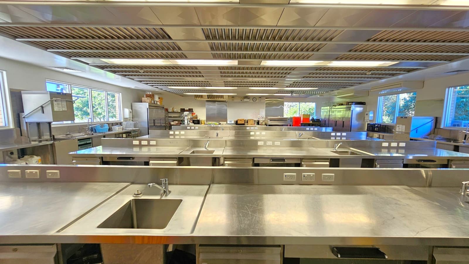 A large, modern kitchen with multiple stainless steel workstations, each equipped with a sink and electrical outlets. The ceiling features metallic ventilation panels. Large windows on the left and right walls allow natural light to illuminate the space. In the background, there are shelves with various kitchen supplies and appliances, including a refrigerator and storage boxes. The floor is not visible.