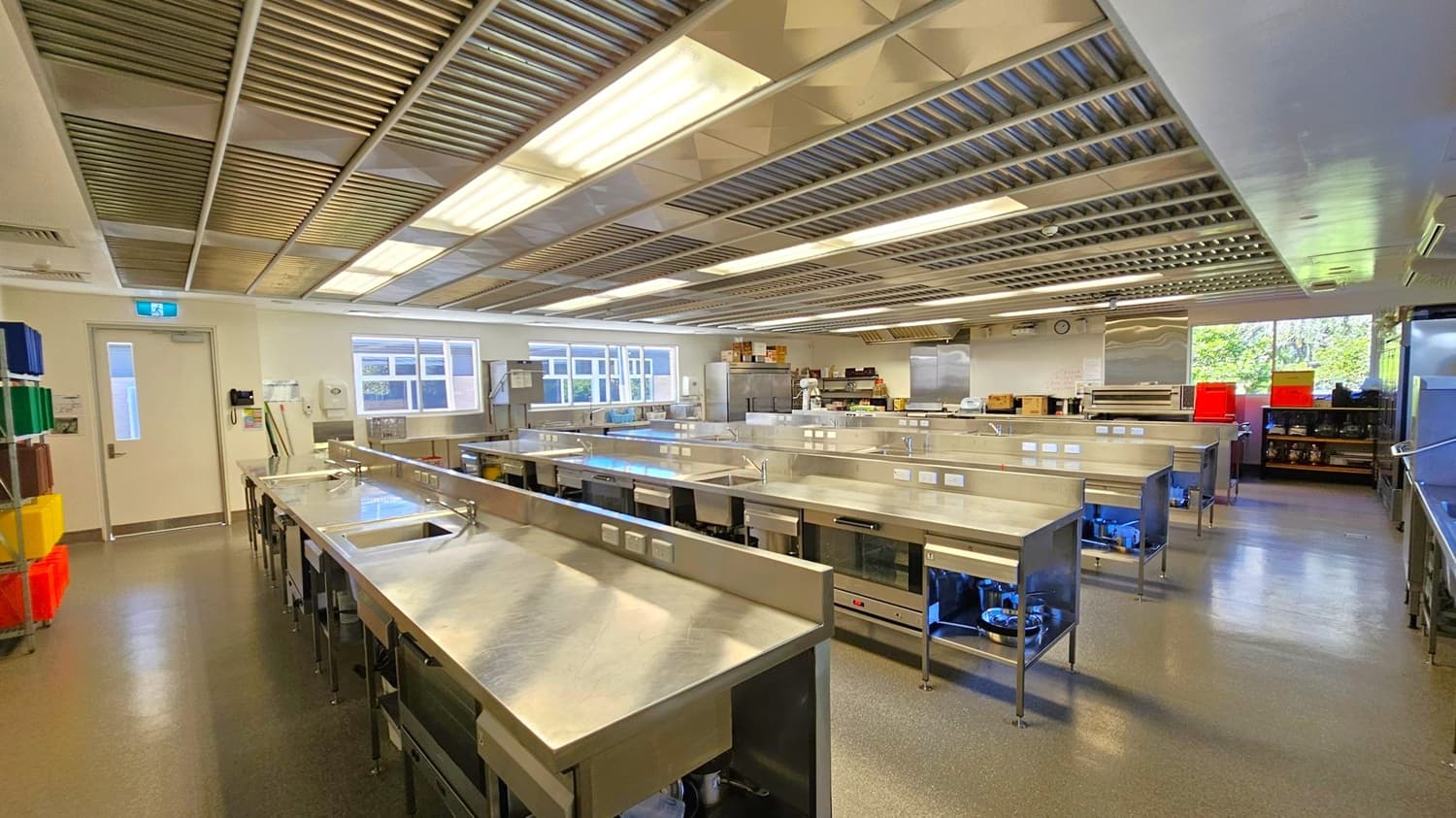 A spacious commercial kitchen with stainless steel workstations arranged in rows. On the left, there are colourful bins stacked on shelves near a white door. The ceiling features metal panels with fluorescent lighting. Large windows on the far wall allow natural light to enter, and there are various kitchen appliances and storage units along the walls. The floor is a smooth, dark material, and the overall space is clean and organised.