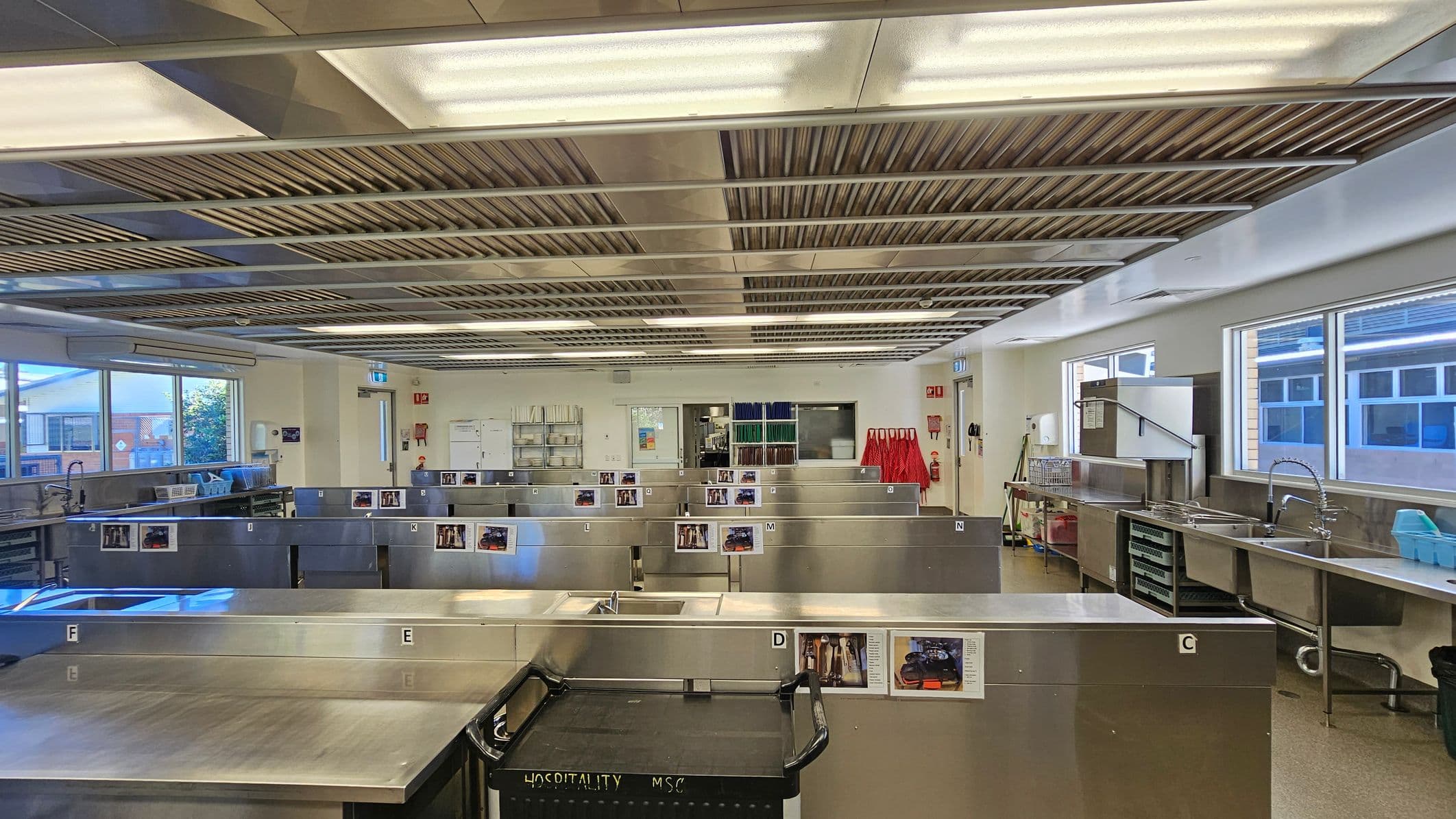 A spacious commercial kitchen with stainless steel workstations arranged in rows. Each station is labelled with letters and has images attached. On the right, there are large windows, a sink, and industrial kitchen equipment. The ceiling features fluorescent lighting and ventilation panels. The floor is a light-coloured material. In the background, shelves with books and red aprons are visible, along with a door and a fire extinguisher.