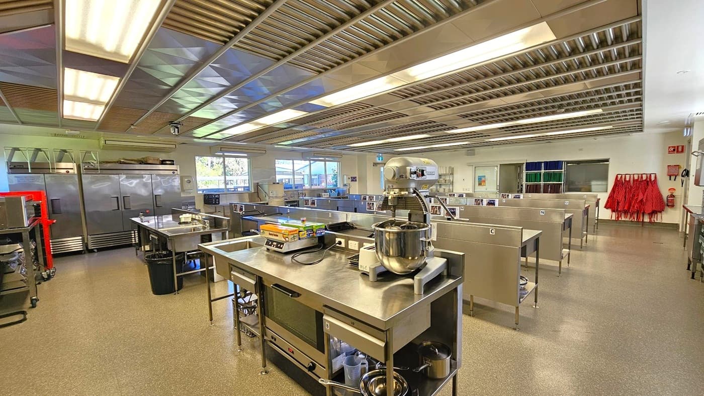 A spacious, modern kitchen with stainless steel workstations and appliances. On the left, there are large refrigerators and storage units. In the centre, a mixer and other kitchen equipment are placed on a metal table. The right side features multiple workstations with sinks and storage. Red aprons hang on the wall near a fire extinguisher. The ceiling has fluorescent lights and a metallic finish, while the floor is a speckled grey. Large windows provide natural light.