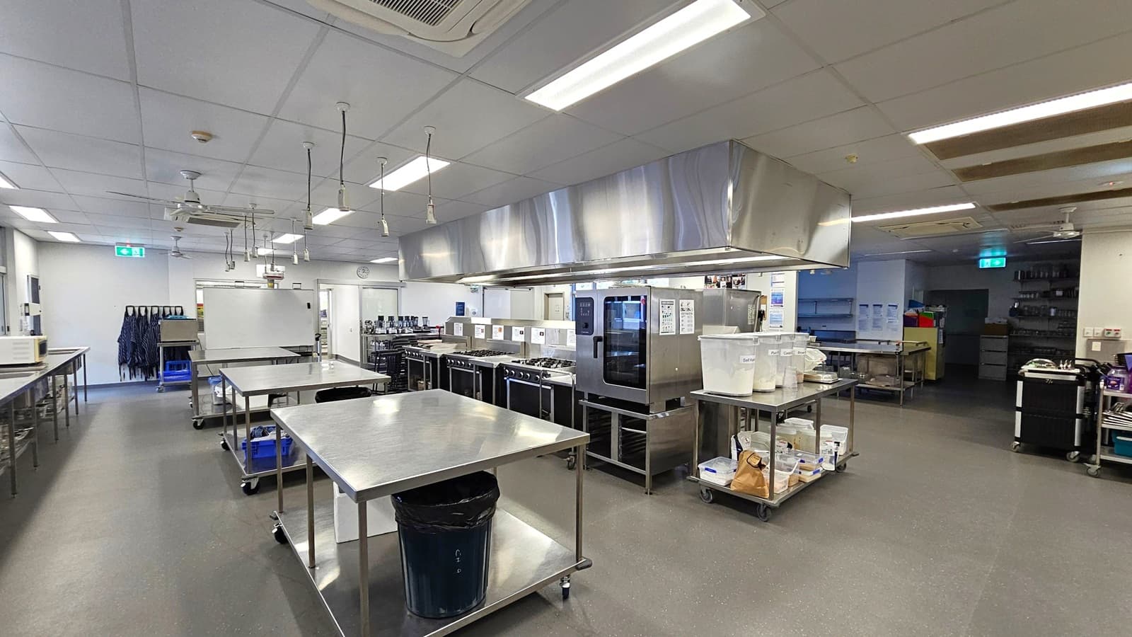 A spacious commercial kitchen with stainless steel worktables in the foreground. On the right, there are large ovens and storage containers. The left side has aprons hanging on the wall and a whiteboard. The ceiling is fitted with fluorescent lights and ventilation ducts. The floor is grey and appears to be made of a durable material. In the background, there are more kitchen appliances and an exit sign above a door.