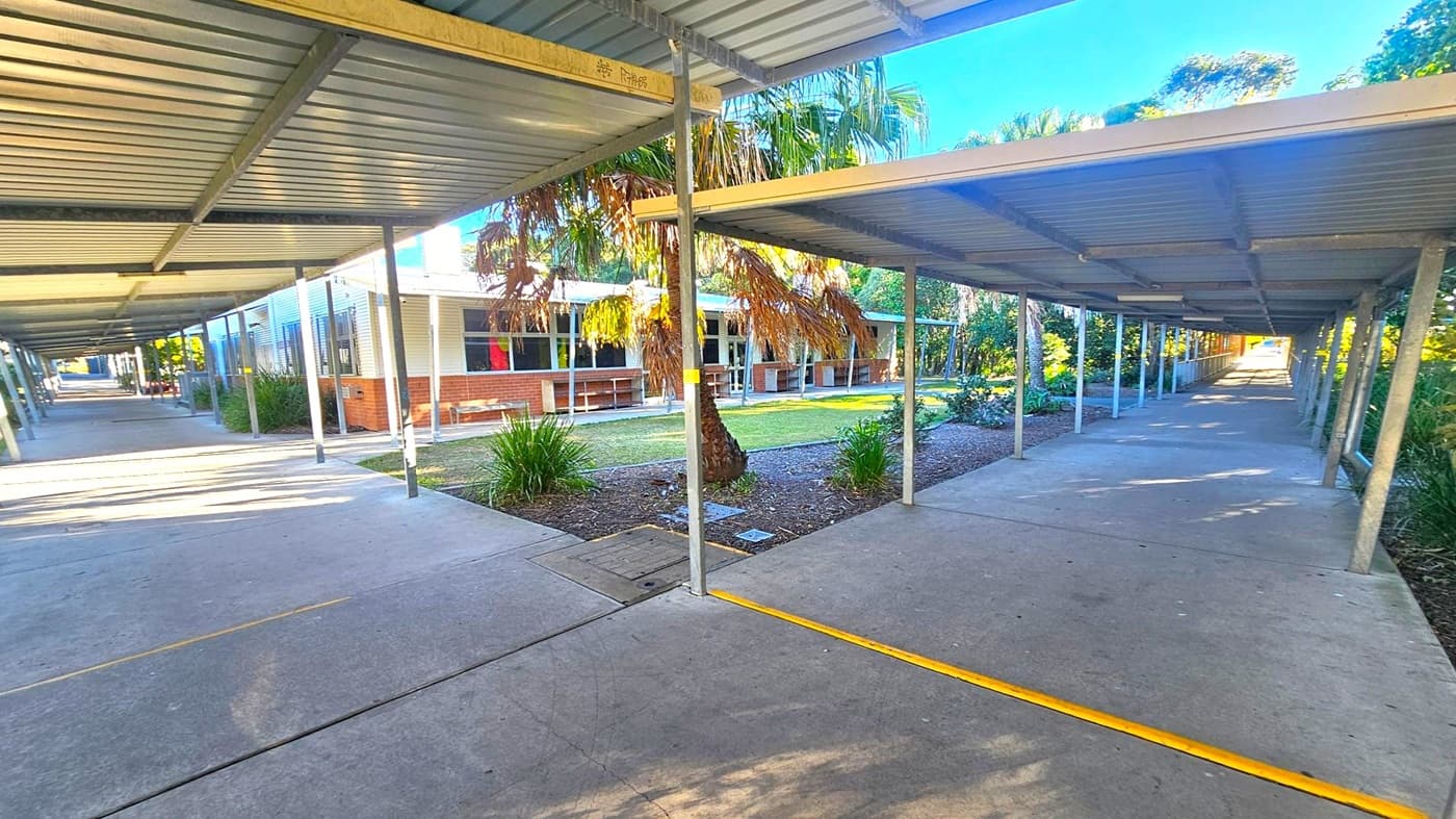 Covered walkways with metal roofs extend into the distance on both the left and right sides. In the centre, a garden area with a palm tree and various plants is visible. A building with brick and white panel walls is on the left, featuring several windows. The ground is concrete, and the area is well-lit with natural light. Trees and greenery are in the background, creating a lush environment.