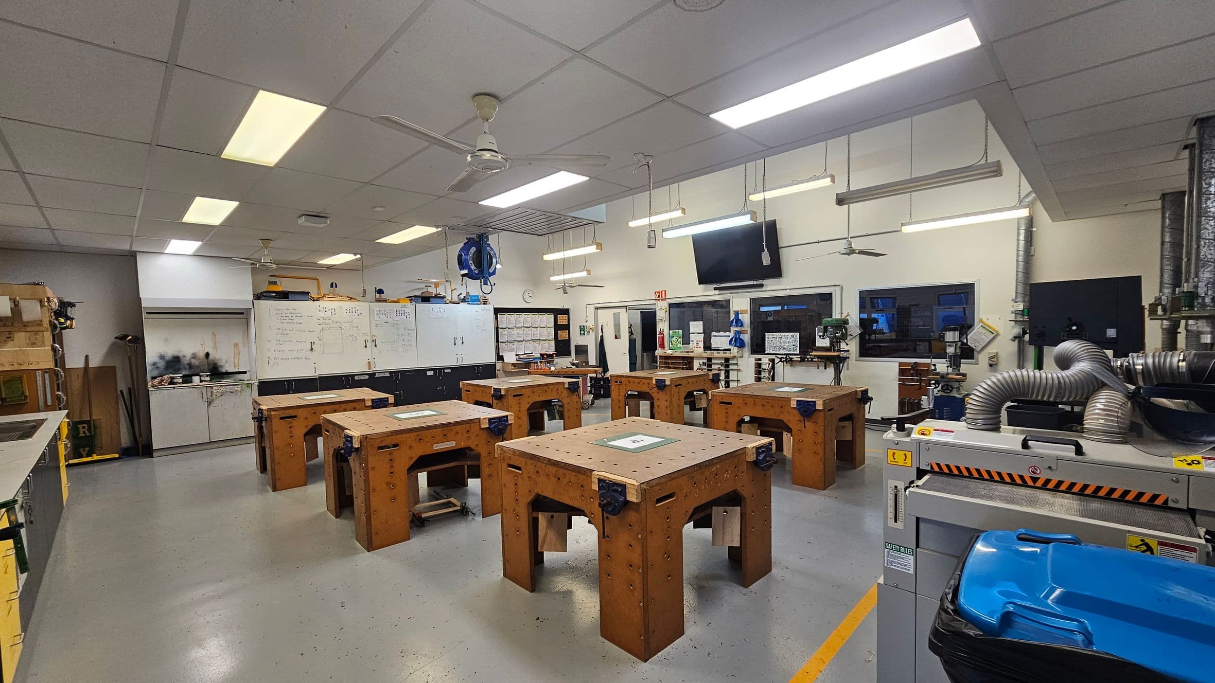 A workshop with a grey tiled floor and a white panelled ceiling. In the centre, there are several wooden workbenches with vices attached. On the left, whiteboards and storage cabinets line the wall. On the right, there are machines with flexible ducting and a blue bin. The back wall features a clock, a wall-mounted TV, and more equipment. Fluorescent lights and a ceiling fan are suspended from the ceiling.