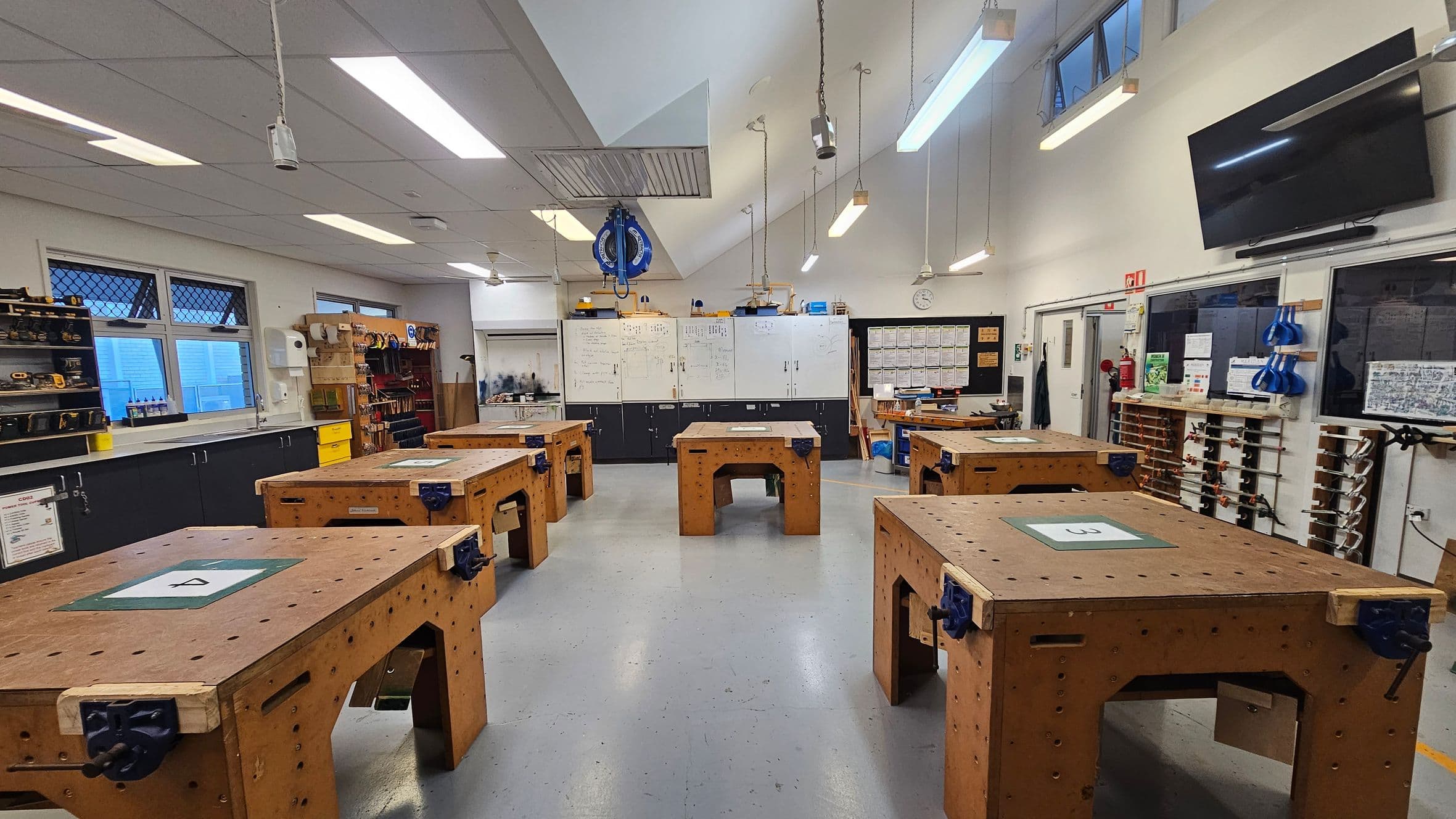 A workshop with several large wooden workbenches arranged in rows. Each bench has a vice attached. The left side features windows with mesh coverings and shelves filled with tools and equipment. The right side has a wall with various tools hanging and a large screen mounted above. The back wall has whiteboards with notes and diagrams. The ceiling is fitted with fluorescent lights, and the floor is a smooth grey surface.