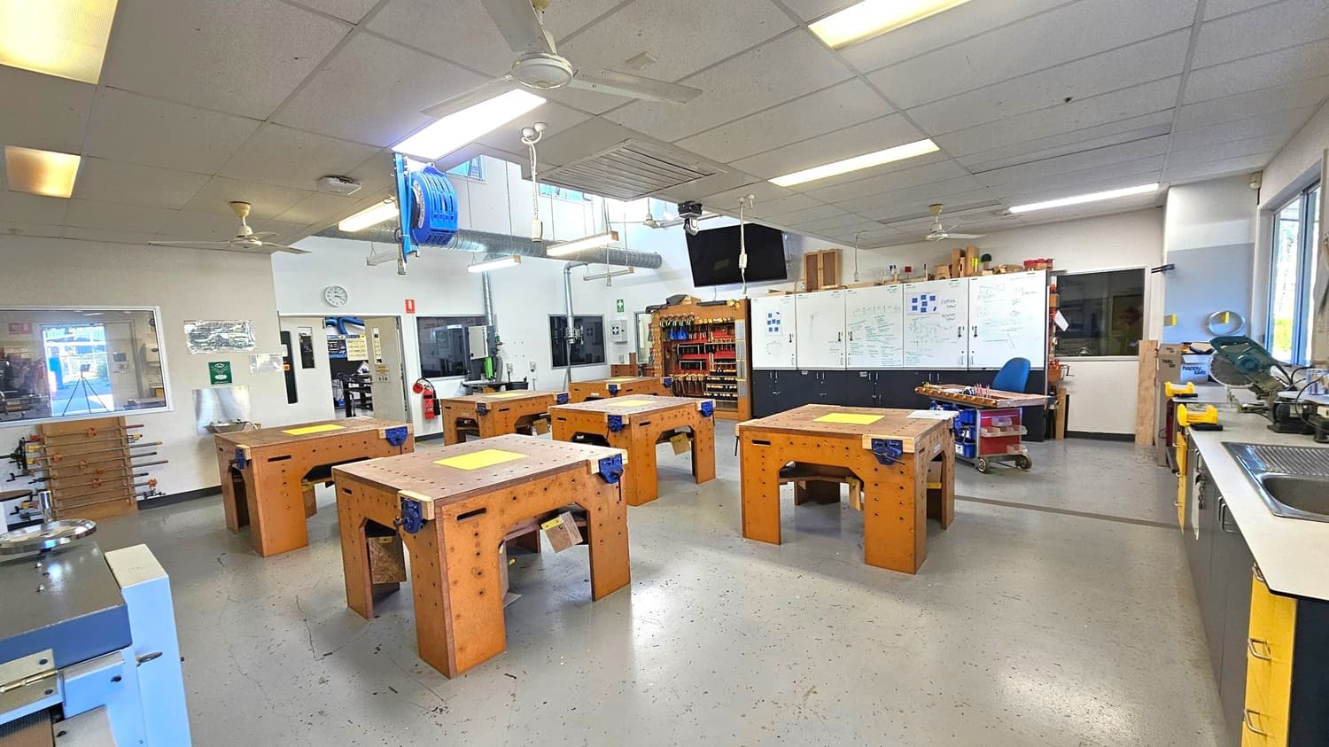 A workshop with several wooden workbenches arranged in the centre, each equipped with a vice. On the right, a long counter with a sink and various tools. The back wall features whiteboards with diagrams and notes, and a rack of tools. On the left, a window and a wall-mounted clock. The ceiling has fluorescent lights and fans, and the floor is grey and speckled. A fire extinguisher is visible near the entrance.