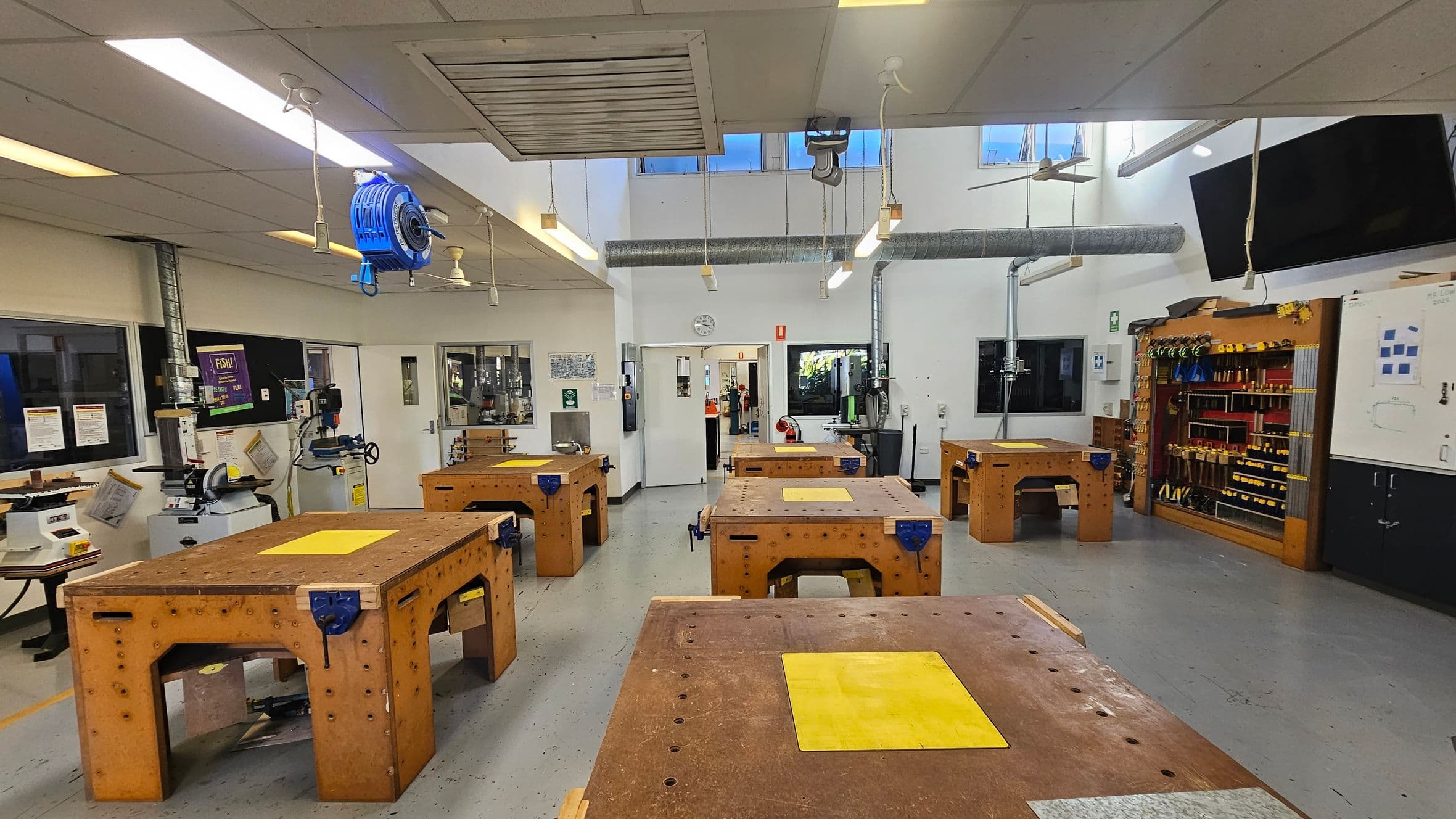 A workshop with several large wooden workbenches, each featuring a yellow mat. On the left, there are various machines and tools near the windows. The right side has a wall-mounted tool rack filled with hand tools. The ceiling has hanging lights and a blue retractable cable reel. The floor is grey and the walls are white, with a clock and safety signs visible. In the background, there is a doorway leading to another room.