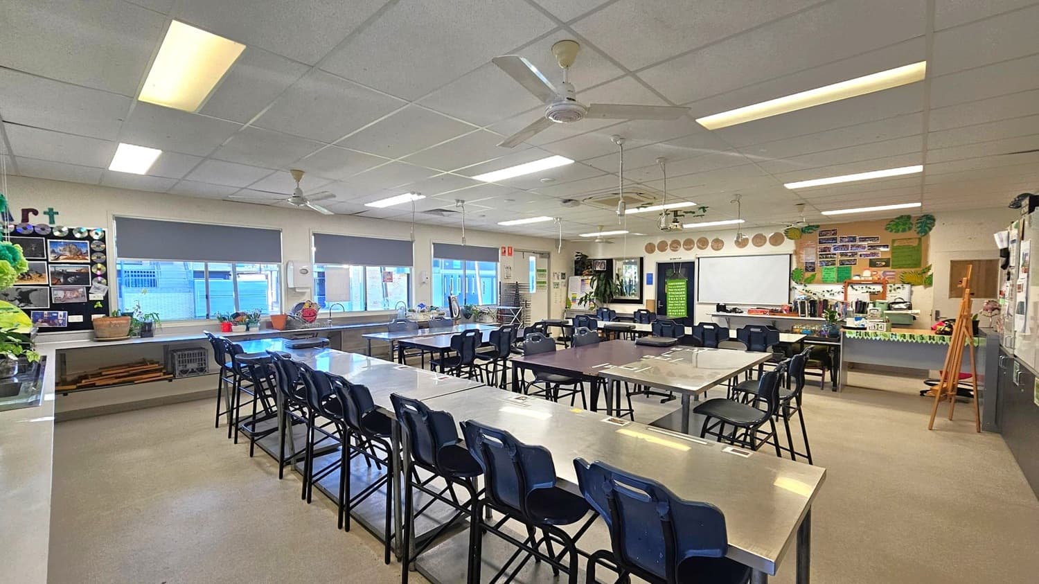 A spacious classroom with several tables and chairs arranged in rows. On the left, there are windows with grey blinds and a display board with various artworks. The right side features a whiteboard and a wall with educational posters. The ceiling has fluorescent lights and ceiling fans. The floor is carpeted, and there are various art supplies and plants around the room.