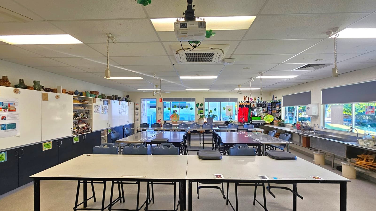A classroom with a white ceiling featuring fluorescent lights and a ceiling-mounted projector. The left side has white cabinets with various art supplies and pottery on top. In the centre, tables and chairs are arranged in rows. The right side has large windows with blinds, a long counter with sinks, and art supplies. The floor is light-coloured, and the walls display art-related posters and decorations.