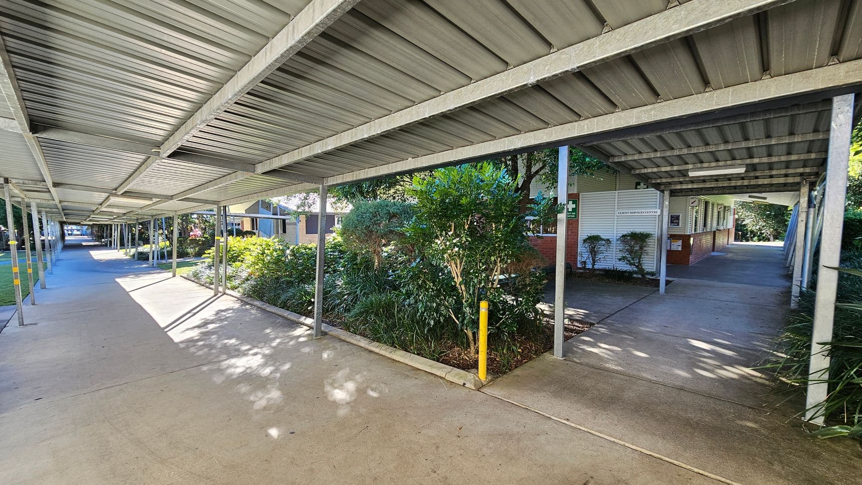 A covered walkway with a corrugated metal roof supported by metal poles extends into the distance. On the right, there is a building with a sign reading "Client Services Centre" and some small shrubs in front. On the left, a garden area with various green plants and trees lines the path. The concrete floor is partially shaded by the roof, with patches of sunlight filtering through.