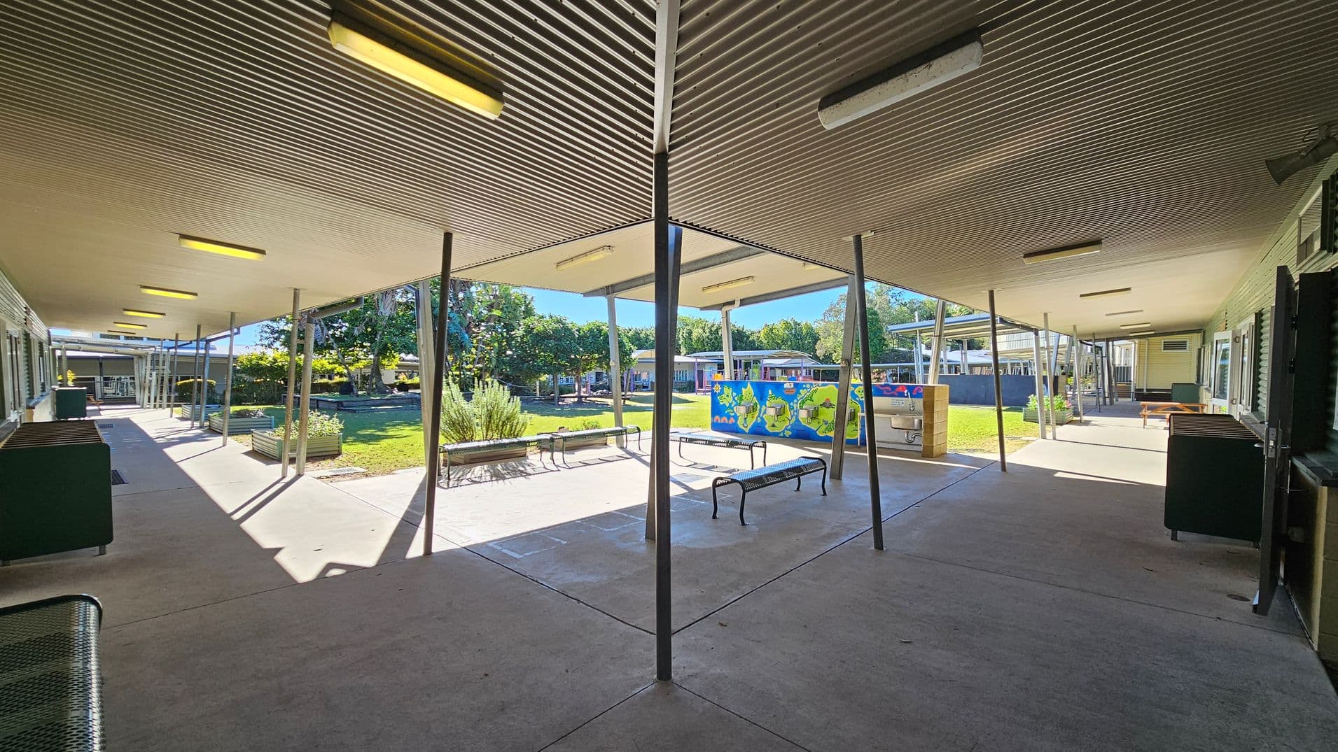 A covered outdoor area with a corrugated metal ceiling and fluorescent lights. The floor is concrete, and there are several metal poles supporting the roof. In the centre, there are benches and a colourful mural on a wall. The background features a grassy area with trees and more buildings. On the left, there are windows and green benches, while on the right, there are more benches and a black cabinet.
