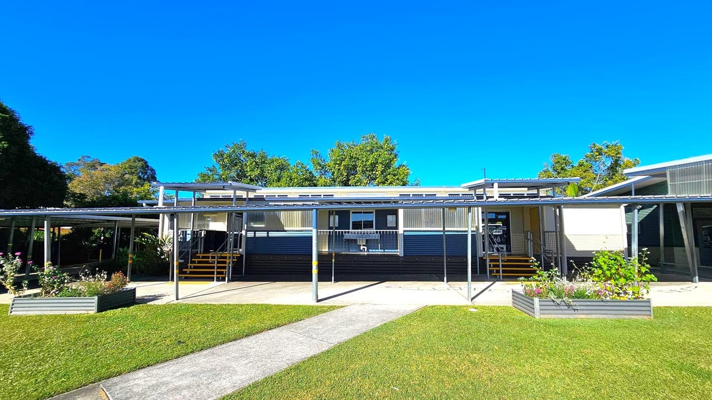 A single-storey building with a corrugated metal roof and a covered walkway supported by metal poles. The building has a central entrance with steps leading up to it. Raised garden beds with flowers are in front of the walkway. The foreground features a well-maintained lawn and a concrete path leading to the entrance. Trees are visible in the background, providing a natural backdrop.