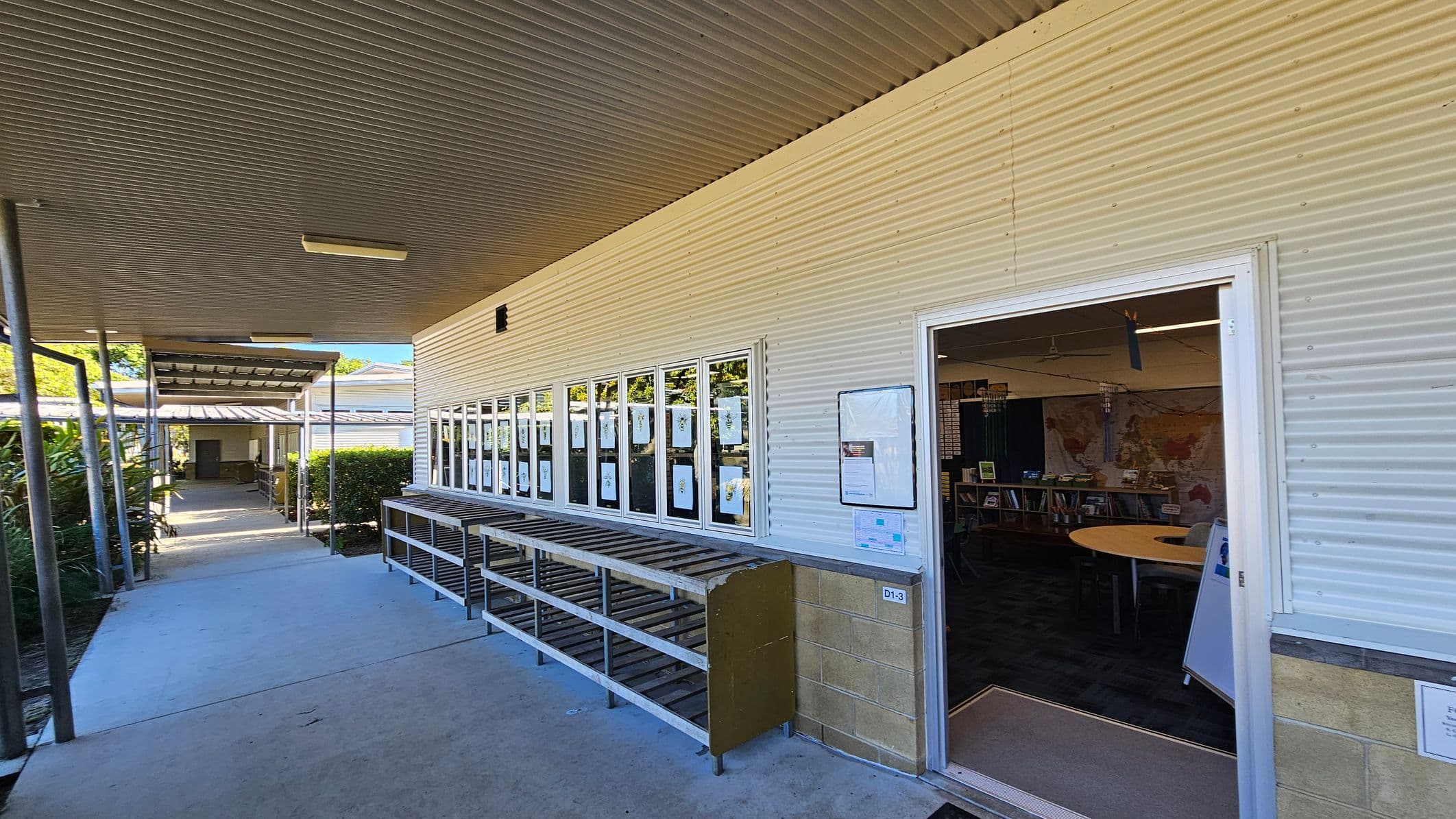 A covered walkway with a corrugated metal ceiling and a concrete floor. On the right, a building with corrugated metal walls features a row of windows displaying papers. An open door leads into a room with bookshelves and a round table visible inside. On the left, the walkway is lined with plants and leads to another building in the distance. A metal shoe rack is positioned beneath the windows.